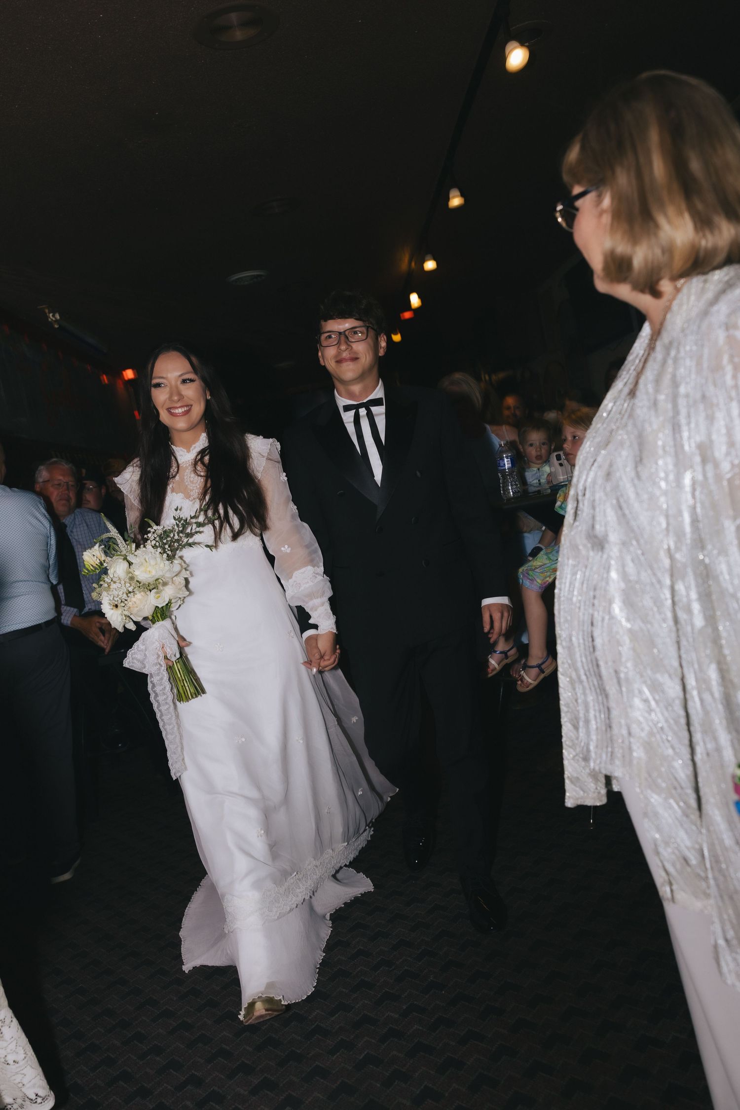 Bride in white gown holding bouquet walks with groom in black tuxedo down wedding aisle.