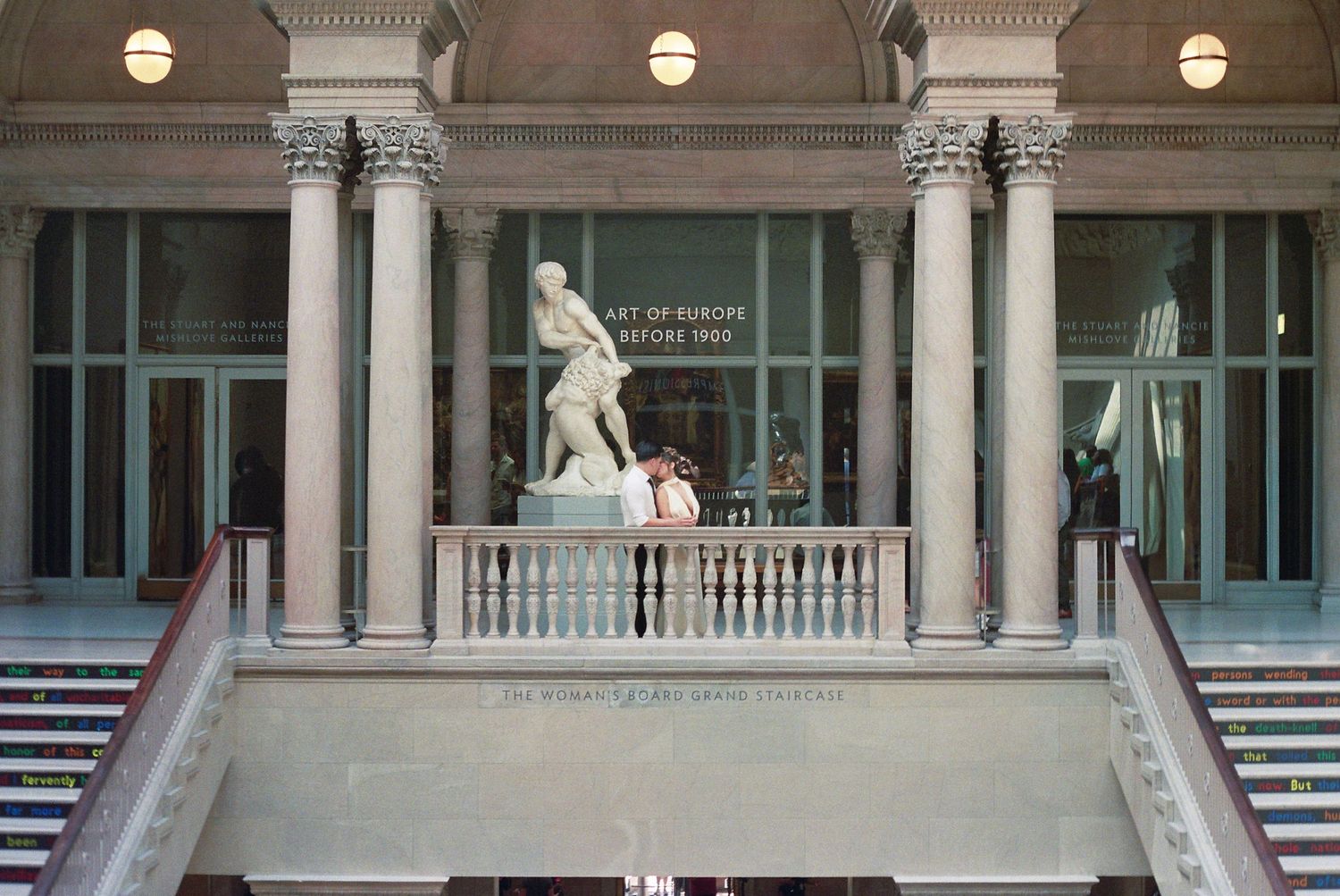 A grand white marble colonnade with ornate columns and round light fixtures mounted along a classical building facade.
