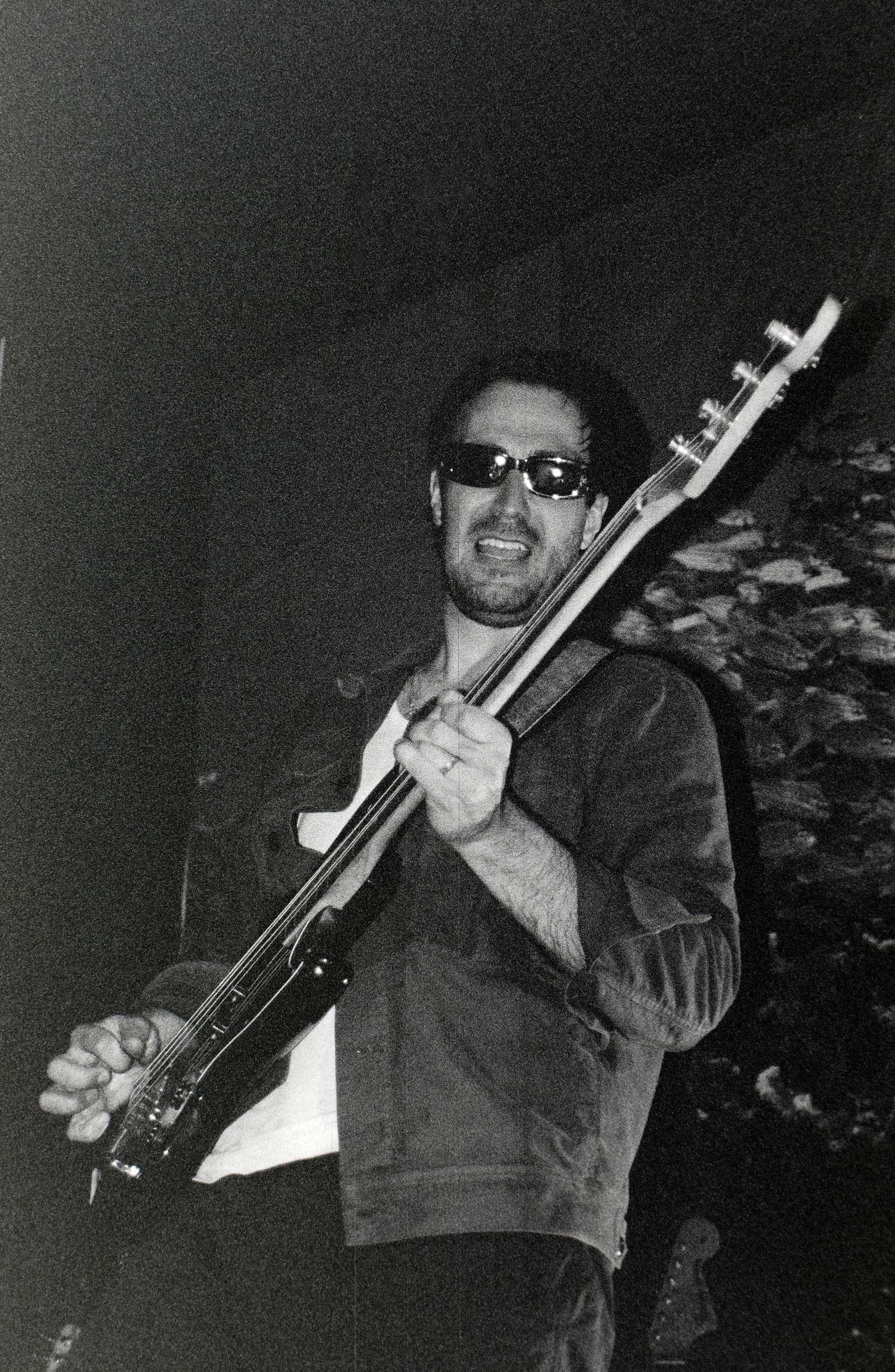Black and white photo of a man playing guitar on stage, wearing sunglasses and a denim jacket.