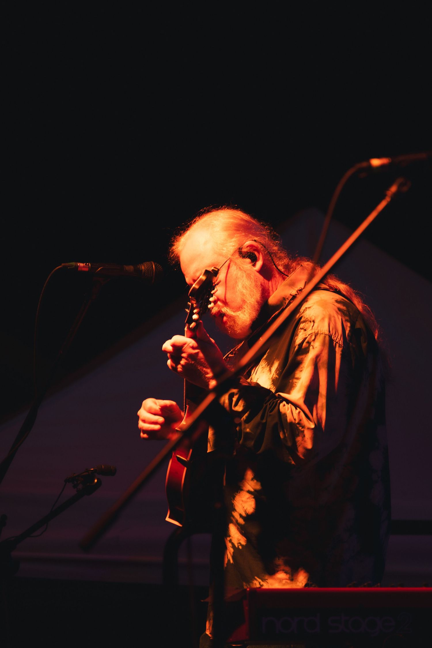 Elderly guitarist performing on stage under warm red lighting with microphone stand in dark concert setting.