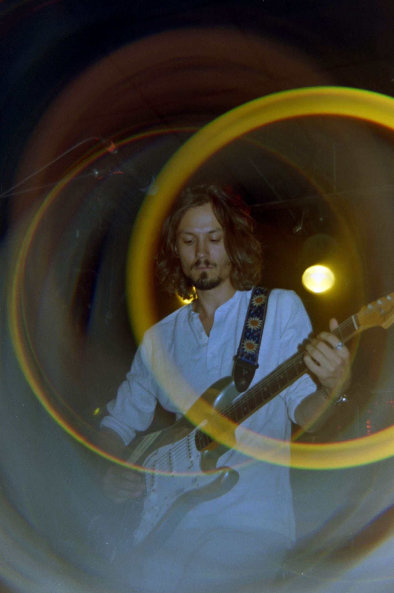 Musician with long hair plays electric guitar on stage, captured with a swirling lens flare effect.