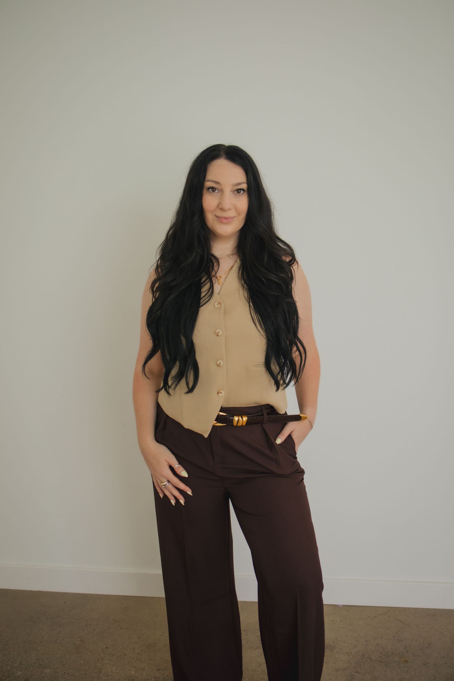 Woman wearing tan sleeveless button-up top and brown wide-leg pants poses against neutral wall.