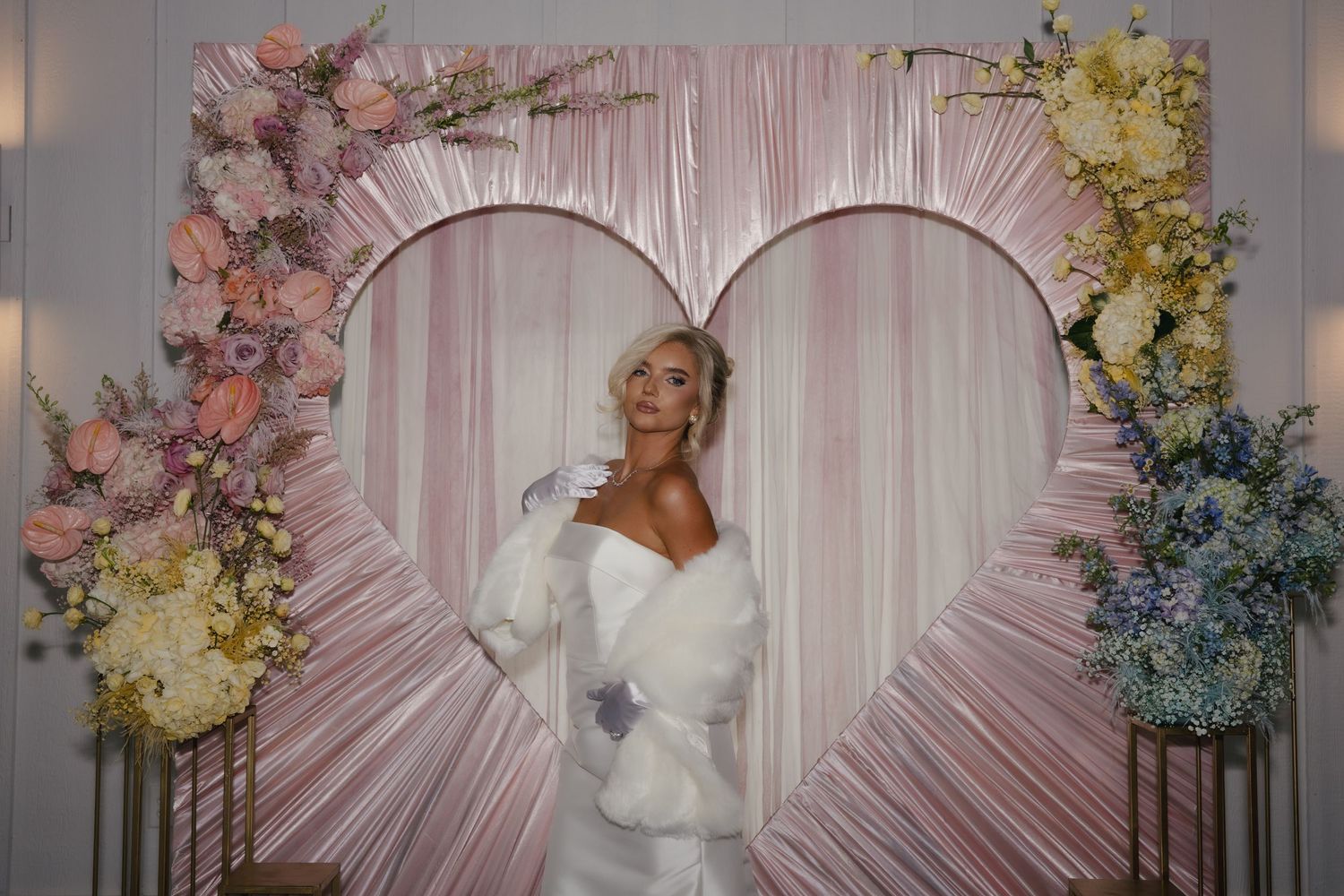 A white-clad figure poses beneath a heart-shaped arch decorated with pink, yellow, and blue floral arrangements.