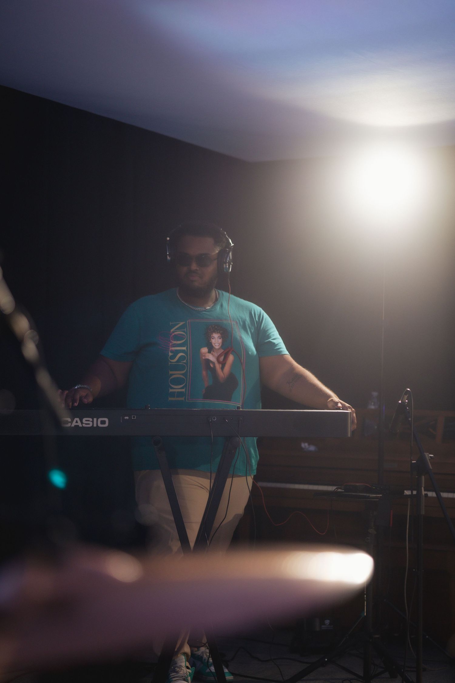 A musician performs on stage with a Casio keyboard in turquoise shirt and headphones under dramatic lighting.