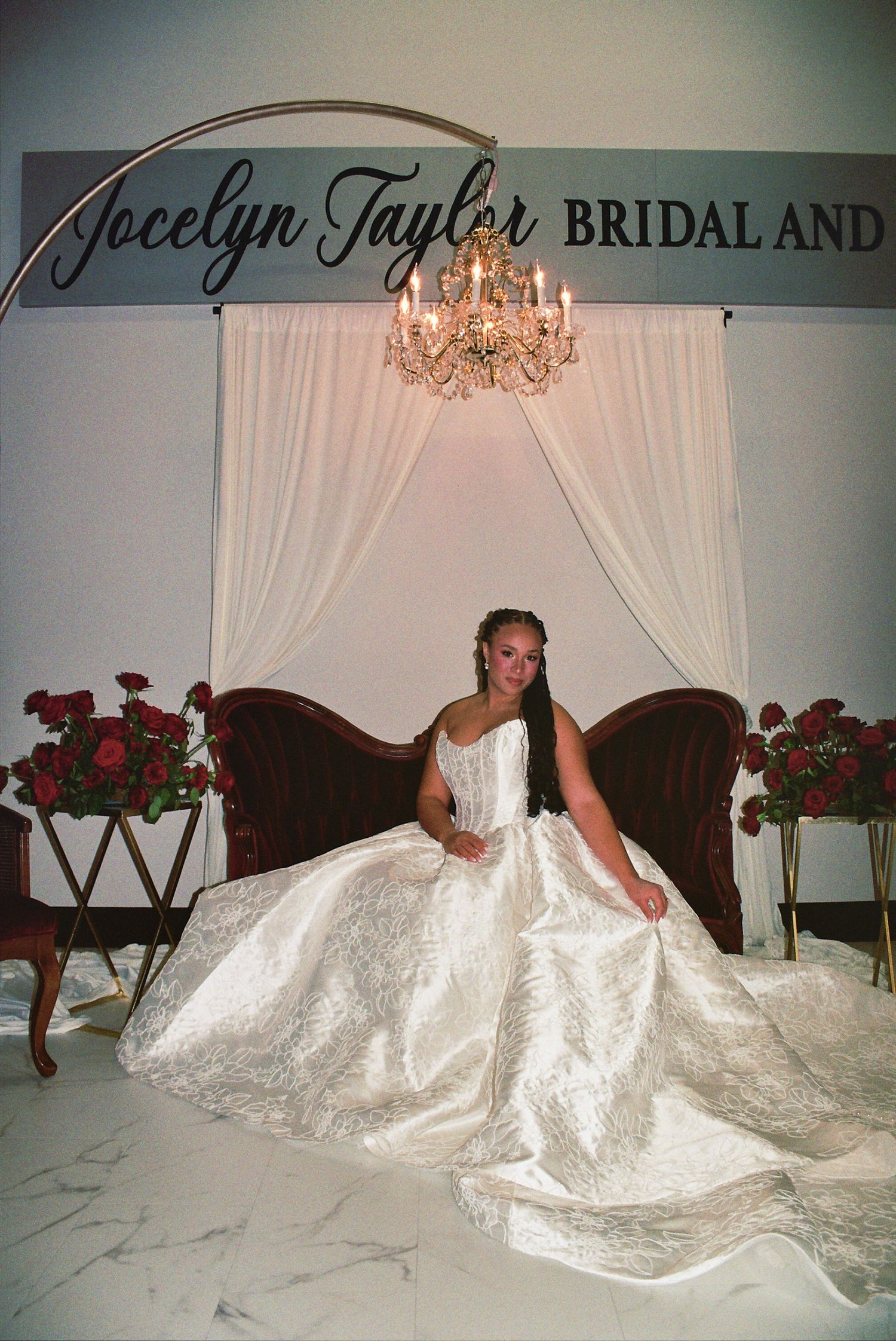 Bride in a voluminous white ball gown seated at Jocelyn Taylor Bridal shop, surrounded by red roses and a chandelier.