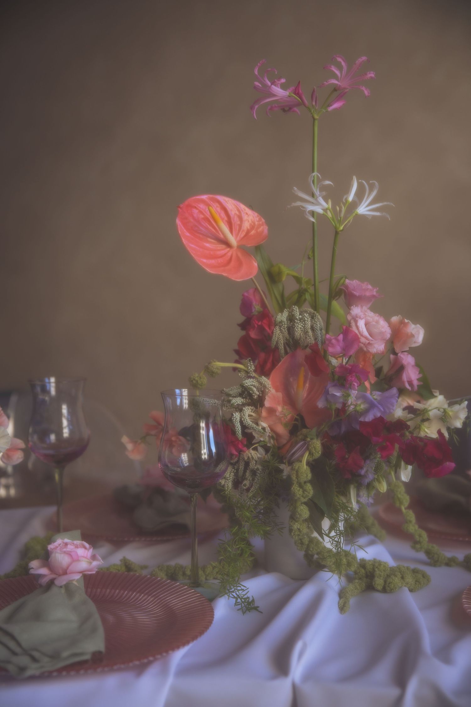 Elegant floral centerpiece with anthurium and pink flowers on white tablecloth with wine glasses creating romantic ambiance.