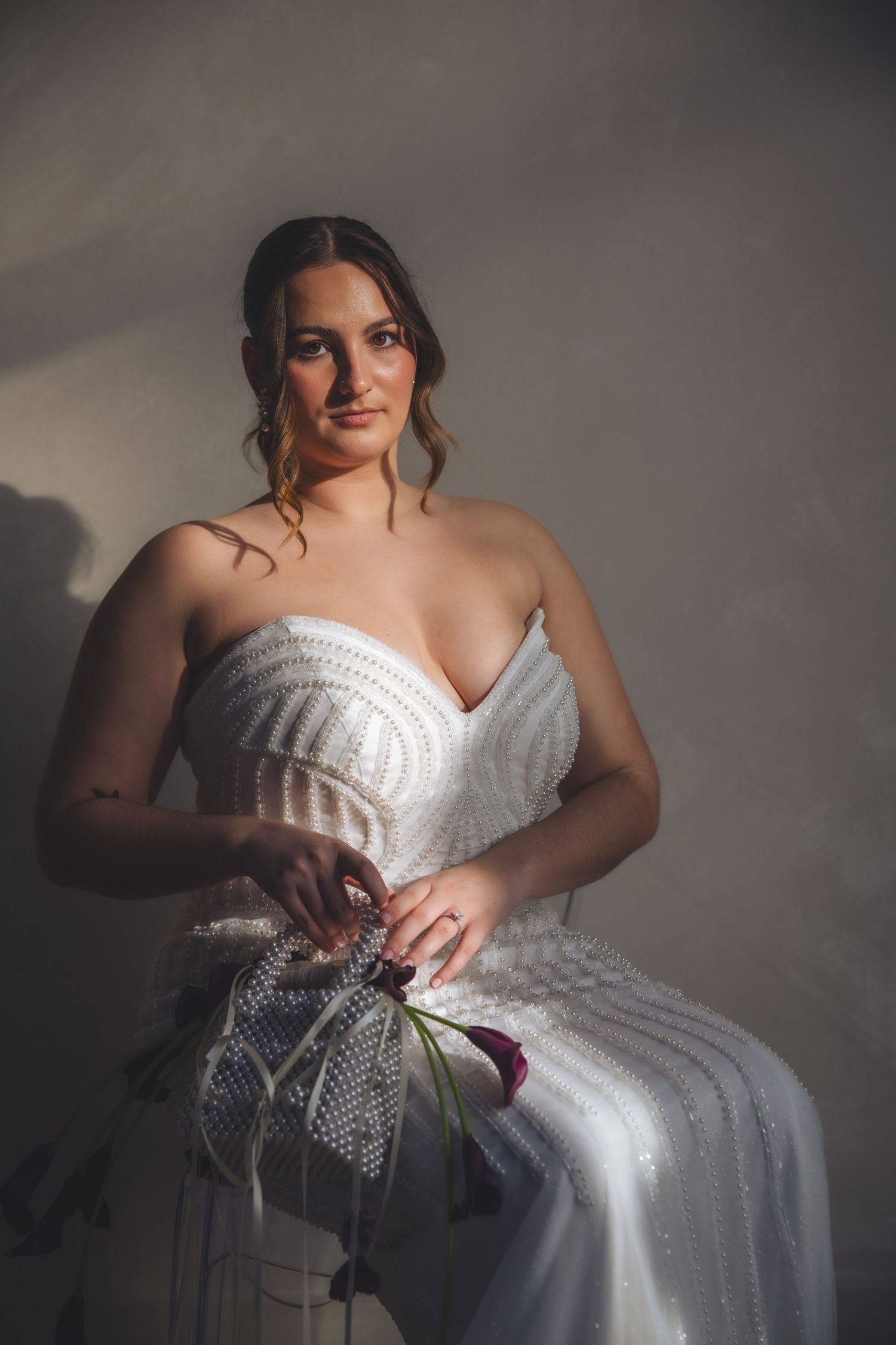 Elegant bride in a strapless white beaded gown poses with bouquet in a moody indoor portrait session.