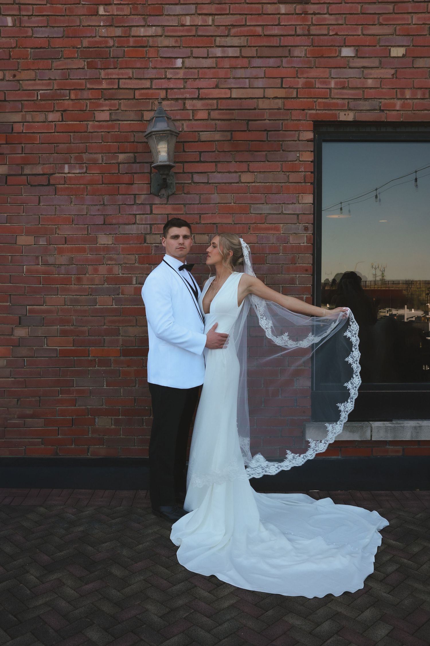 A wedding couple poses against a brick wall at sunset with the bride's delicate lace veil flowing in the breeze.