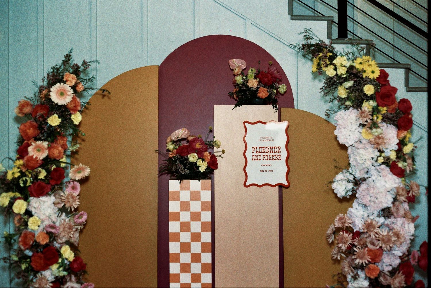 Decorative floral arch with burgundy and cream flowers featuring a checkerboard pattern and event signage.