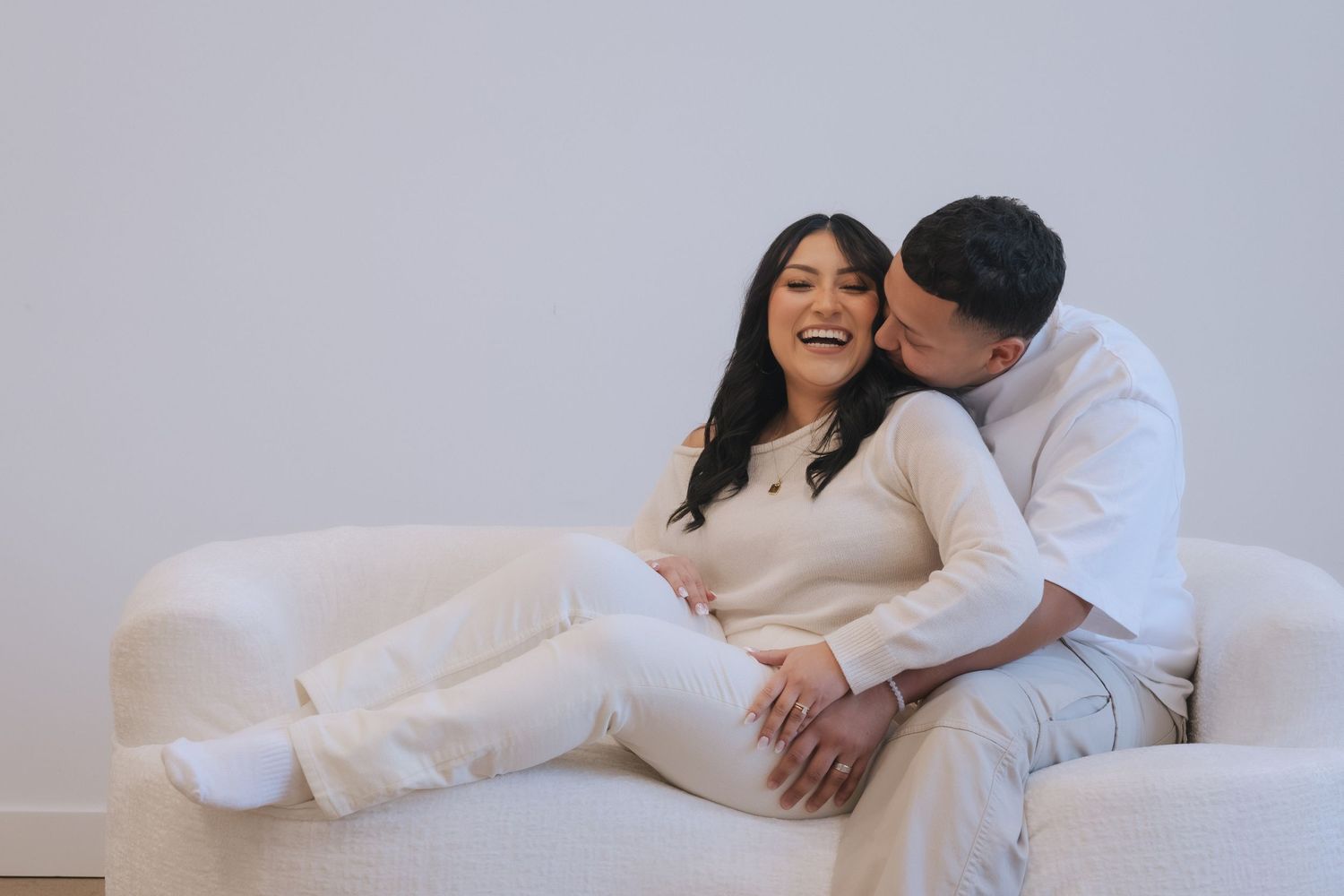 Couple sitting on white sofa, woman laughing as man kisses her cheek, both wearing white outfits.