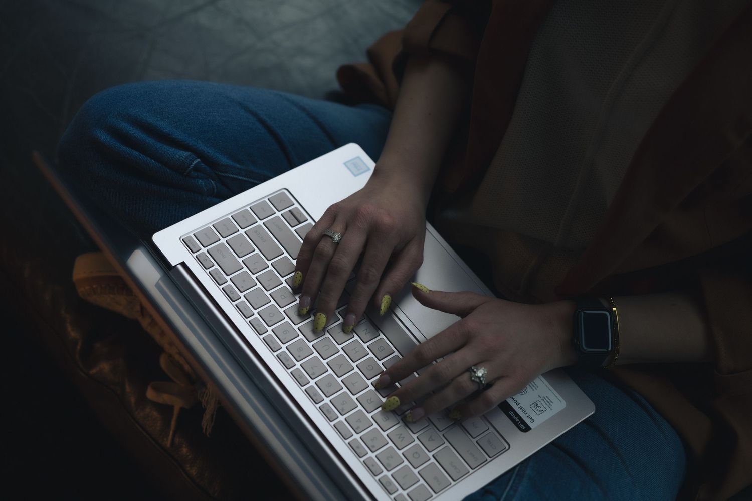 Person typing on a silver laptop keyboard while sitting cross-legged, wearing a smartwatch and rings.