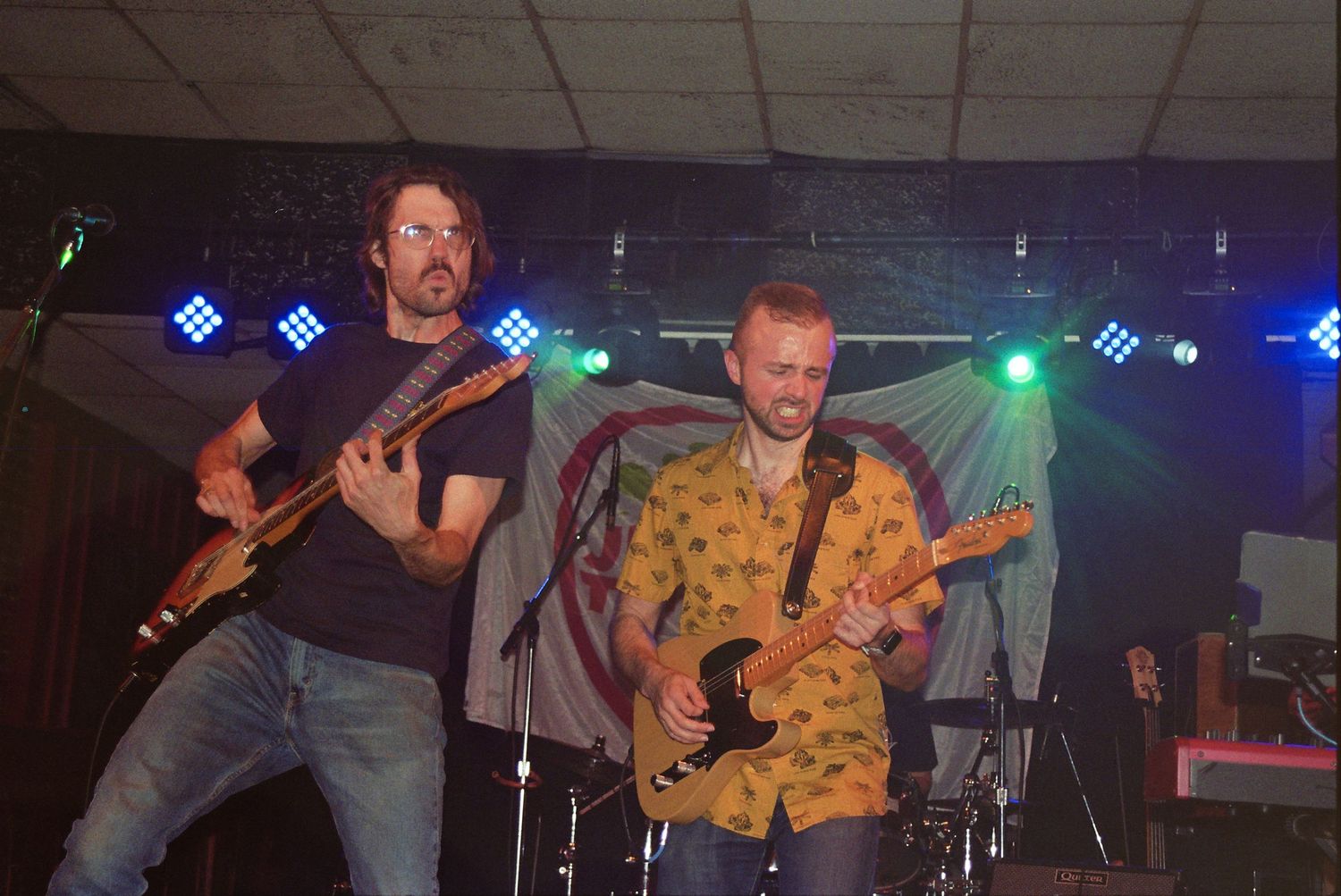 Two male guitarists performing on stage with colorful lights and a banner backdrop at a live music event.