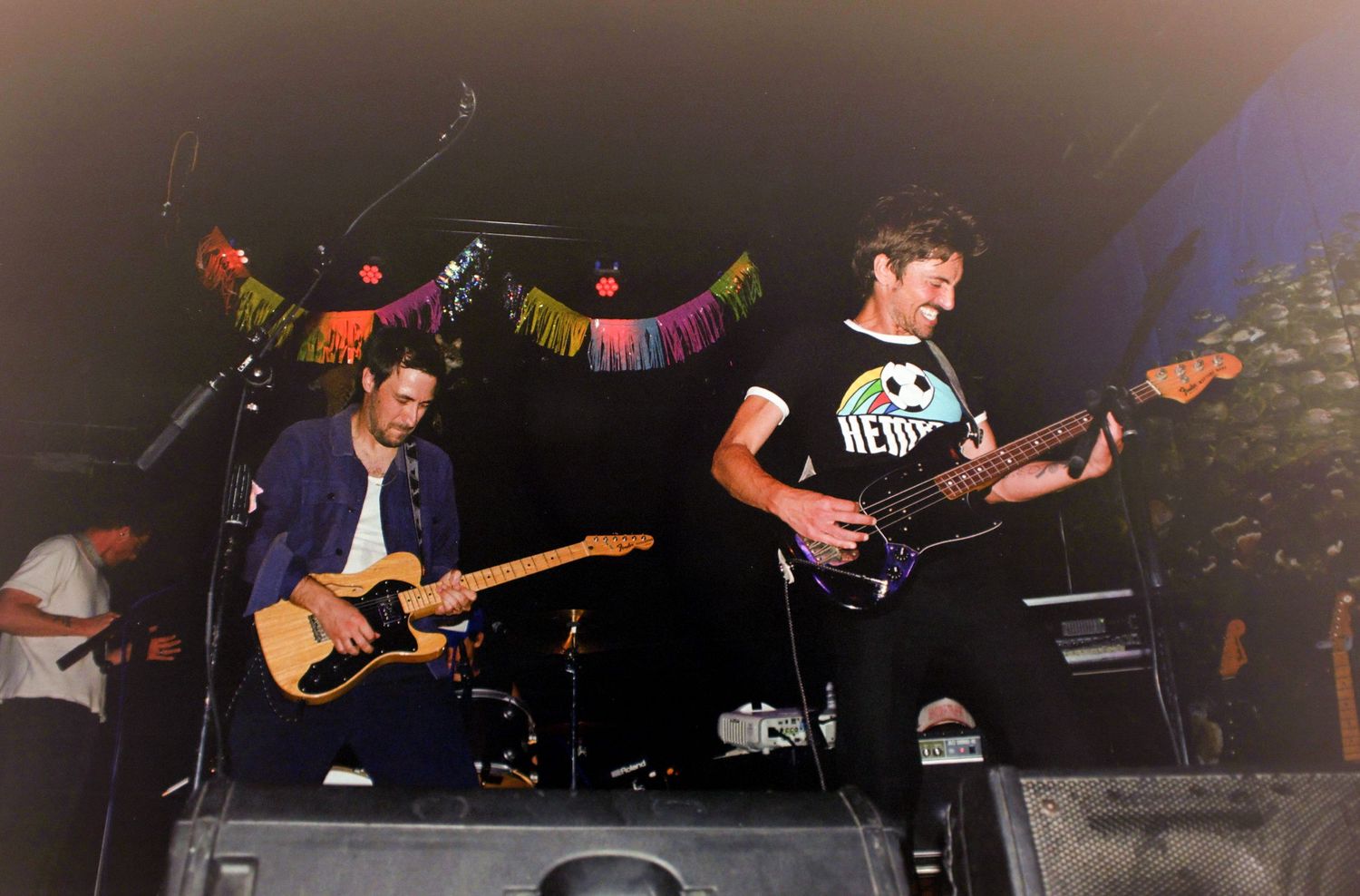 Two guitarists performing on stage at a live music event with colorful bunting decorations in the background.