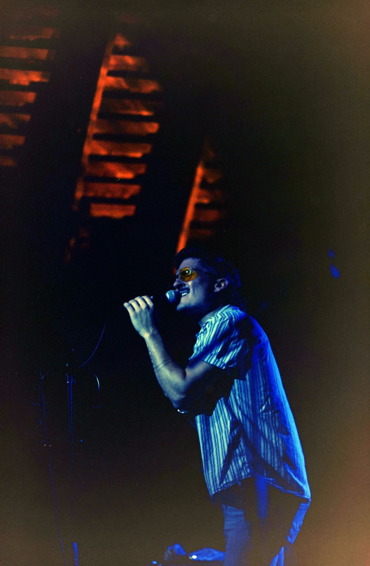 A male singer performs on stage under blue lighting, holding a microphone while wearing a striped shirt.