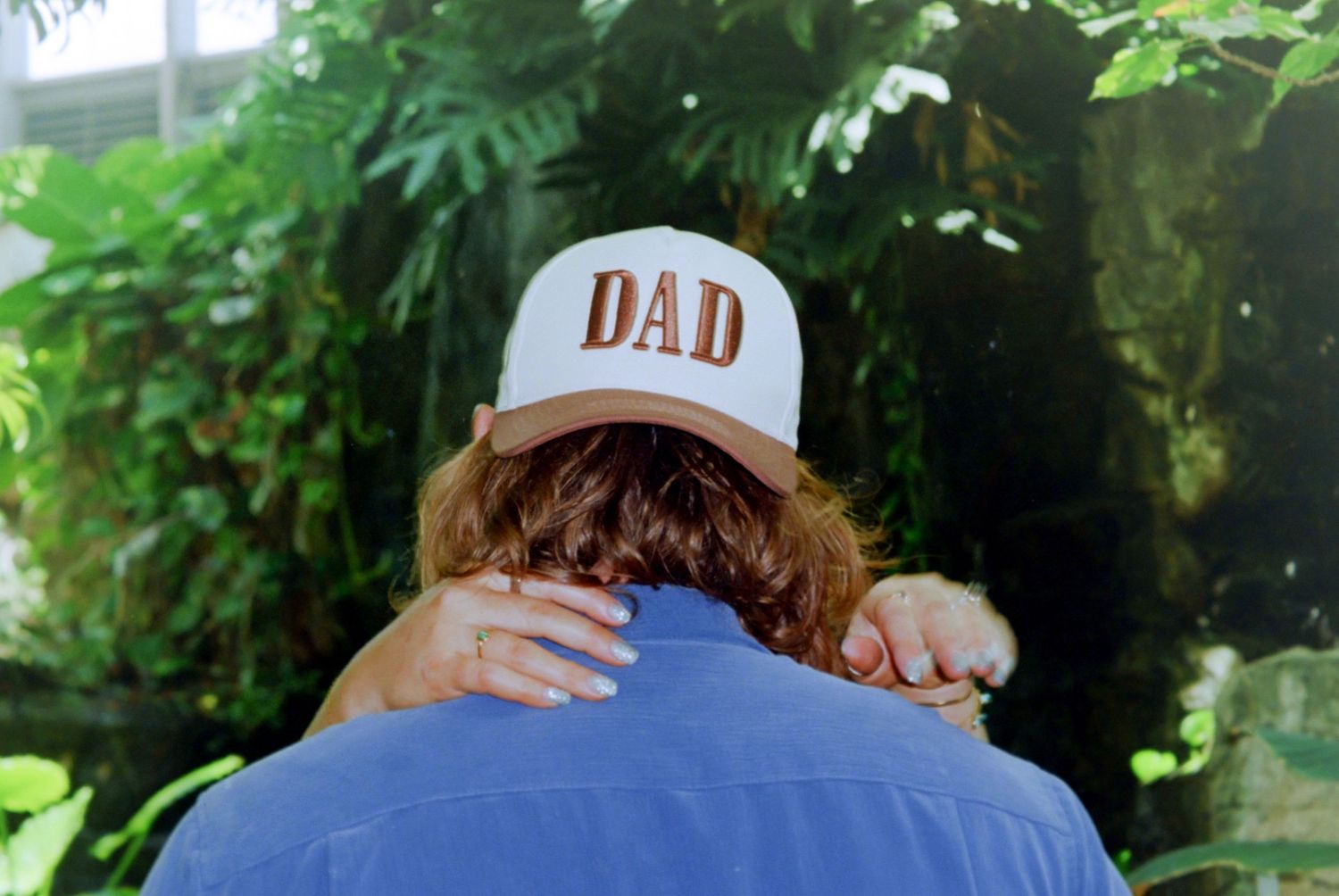 Person wearing a white DAD trucker hat embracing someone outdoors surrounded by lush green tropical foliage.