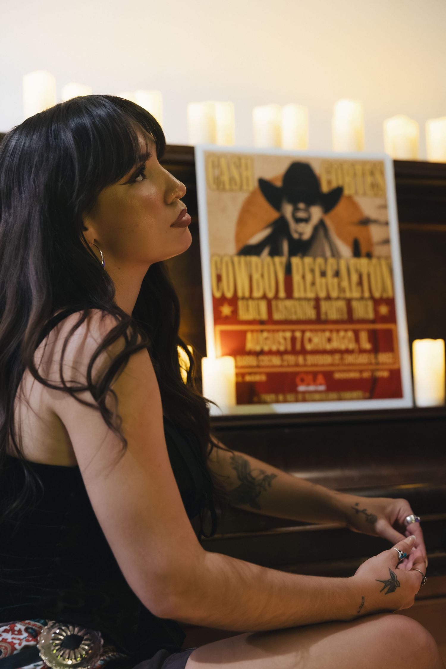 Woman with tattoos sits near candles and a Cowboy Reggaeton Chicago event poster in a moody setting.