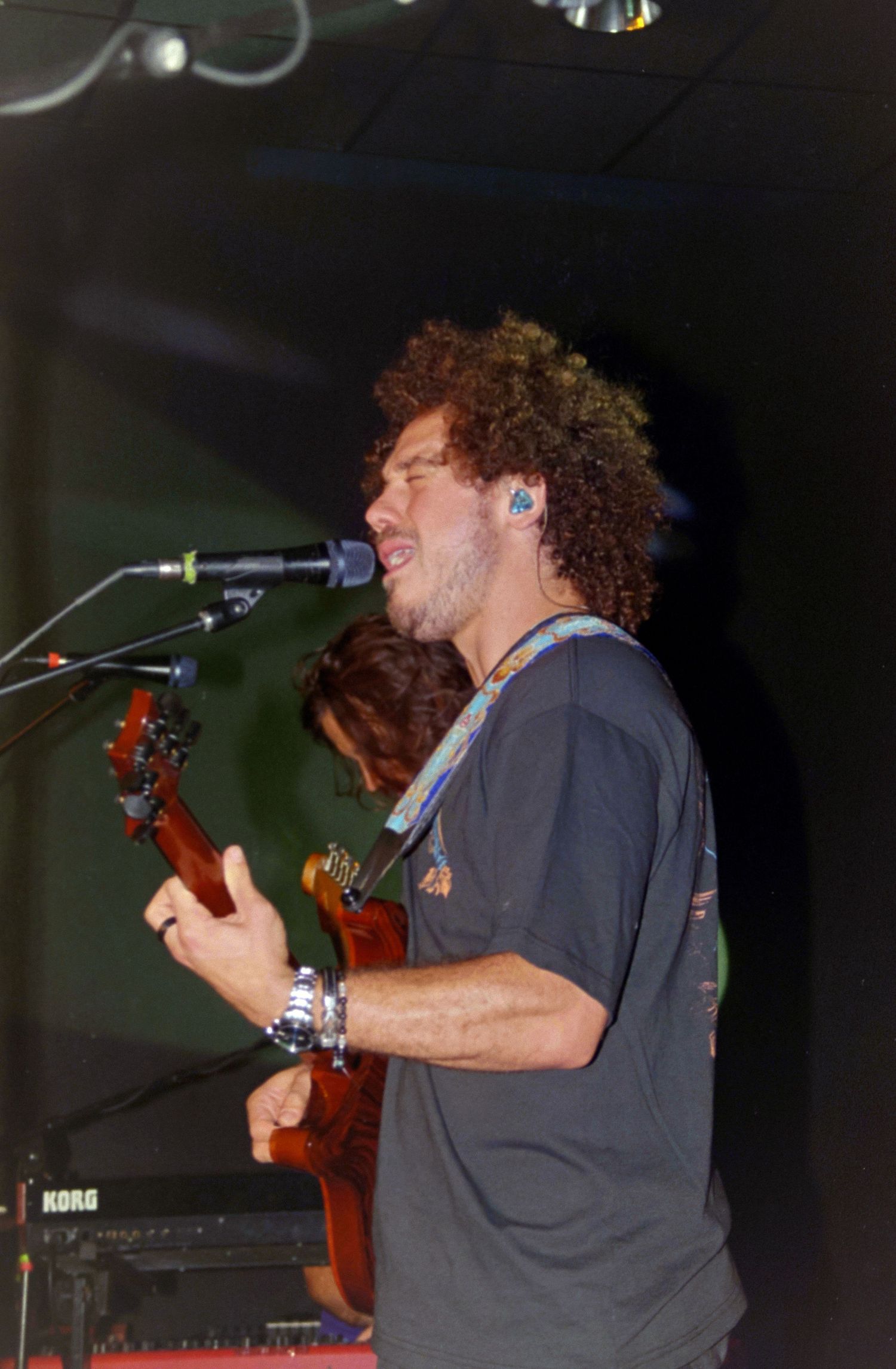Curly-haired male guitarist singing into microphone on stage, playing electric guitar at live concert with keyboard visible.