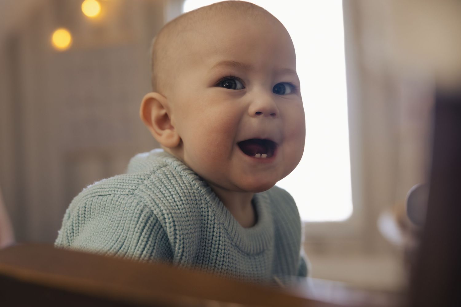 A baby in a gray sweater laughs joyfully while sitting at a table with blurred lights in the background.