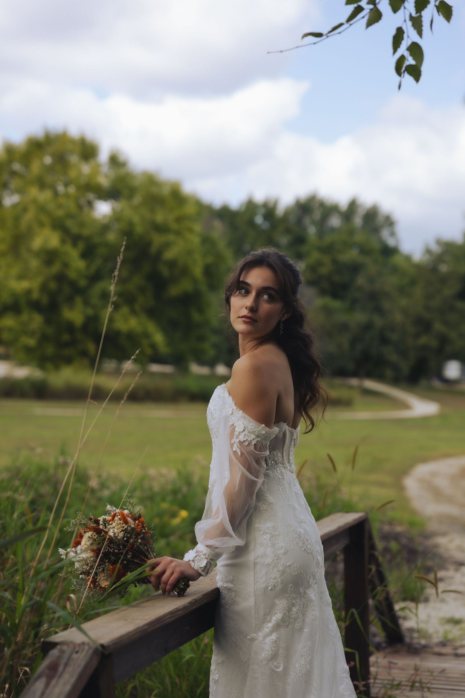 A person in a white dress poses thoughtfully on a wooden railing along a path with green trees in the background.