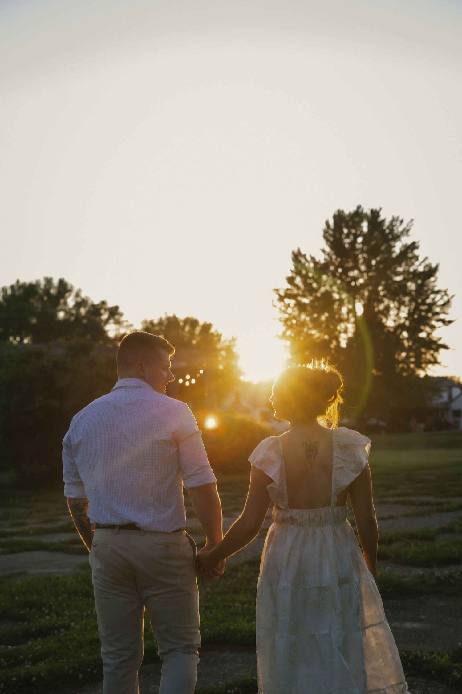 A romantic couple holding hands watching the sunset in a grassy field with golden light filtering through trees.