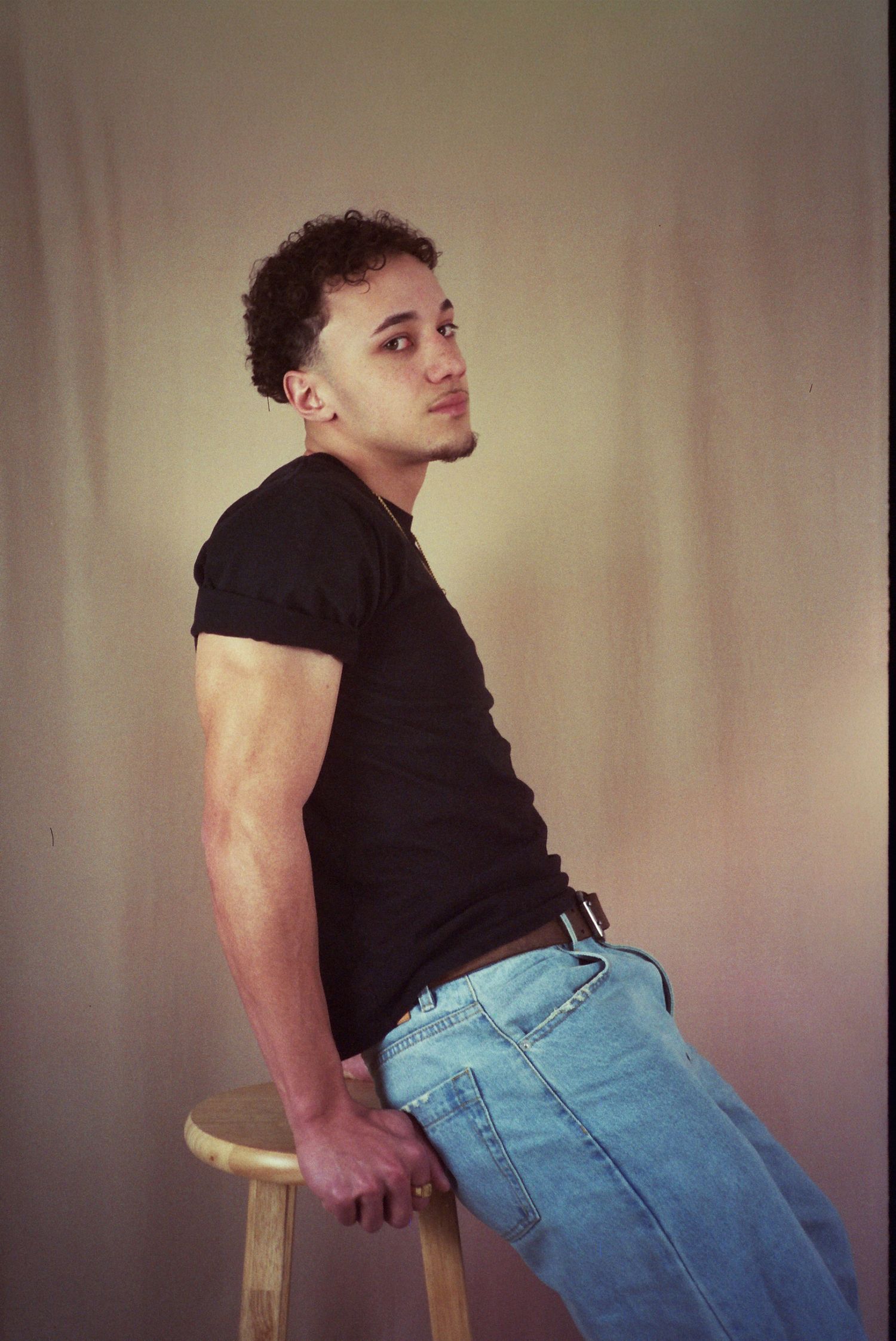 Young man in black t-shirt and light blue jeans sitting on a wooden stool against a neutral background.