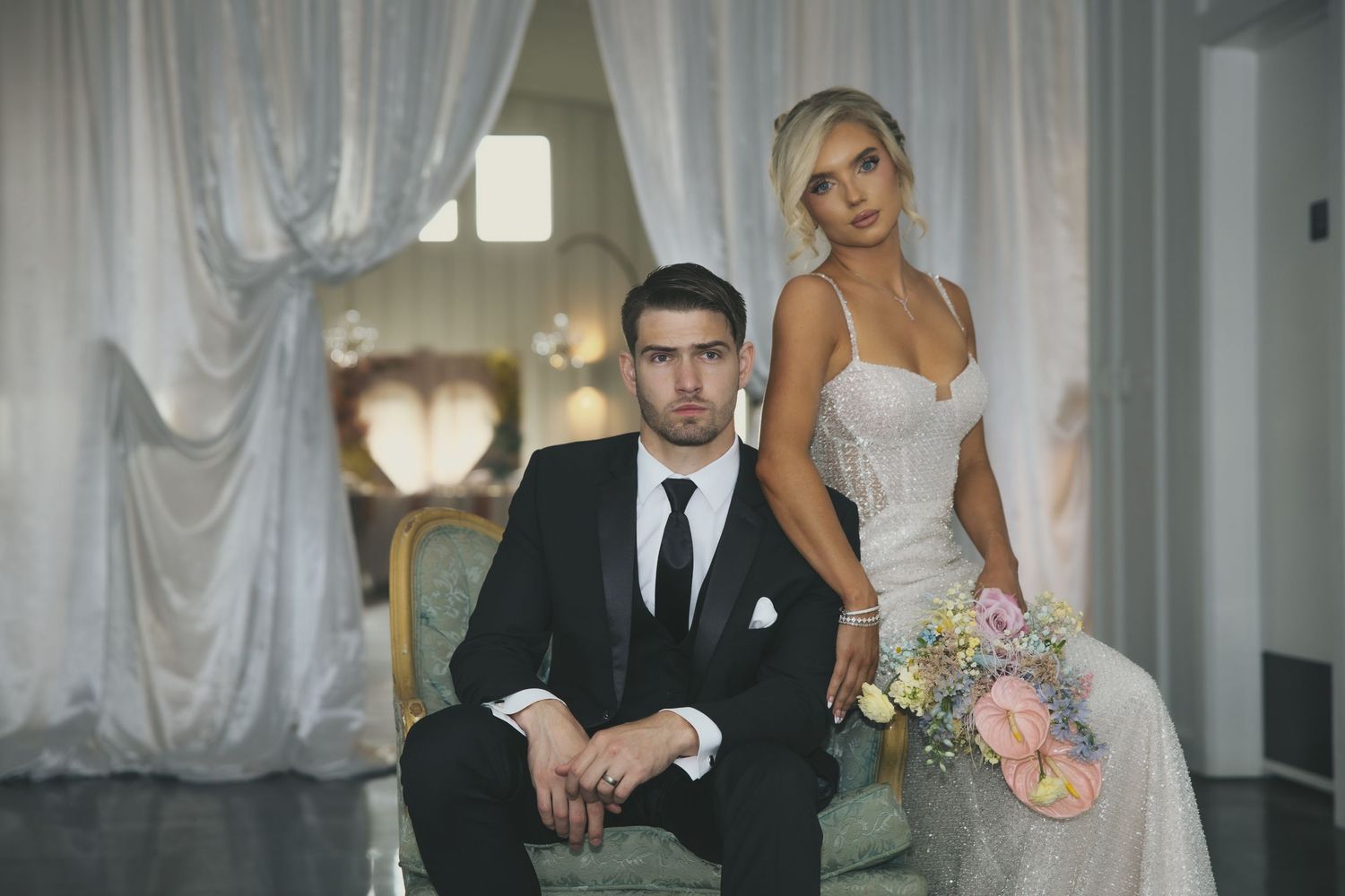 Elegant couple poses in formal wedding attire in front of white curtains with soft lighting.