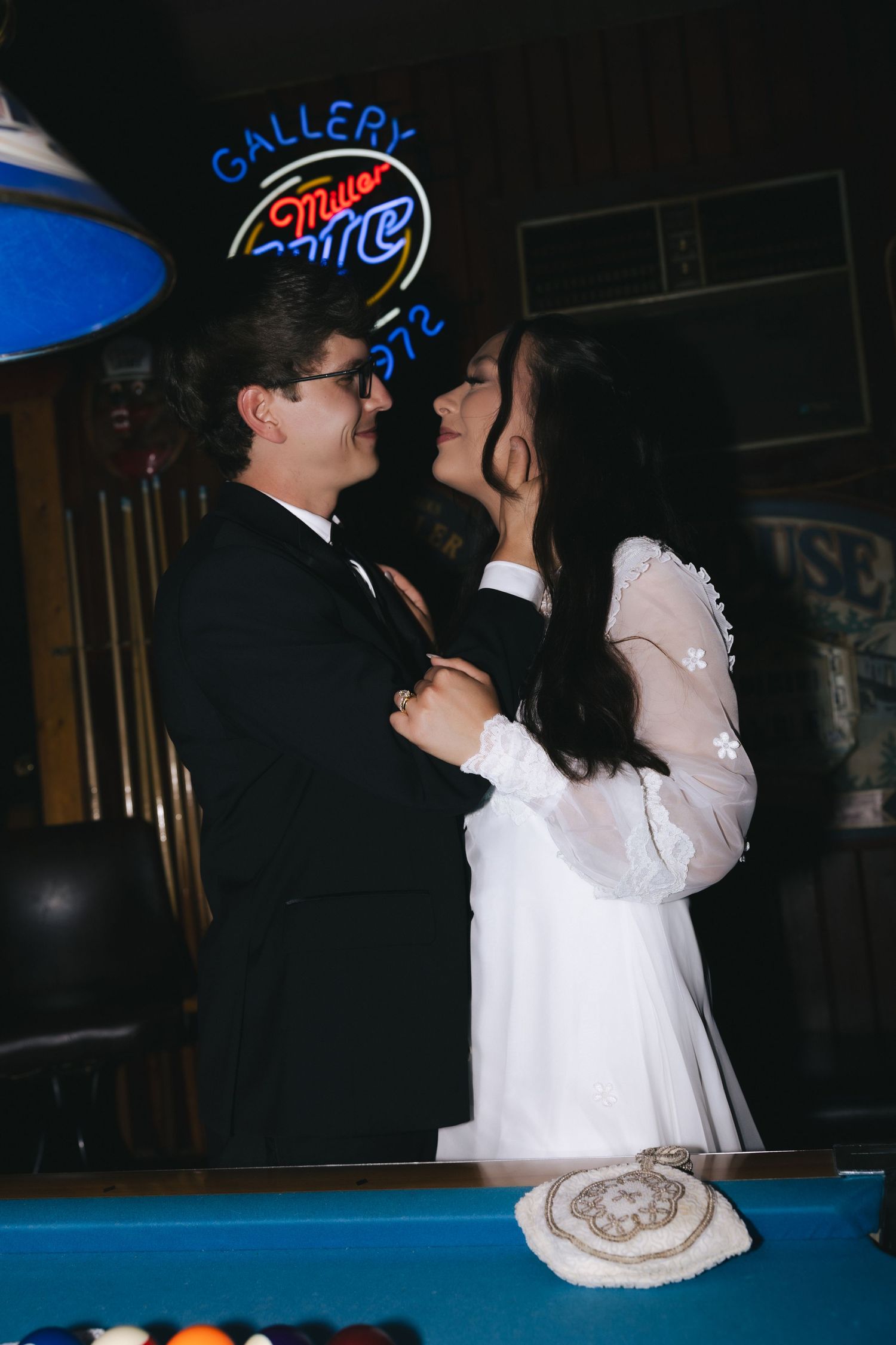 Couple in wedding attire sharing a romantic moment near a pool table under neon bar signs.