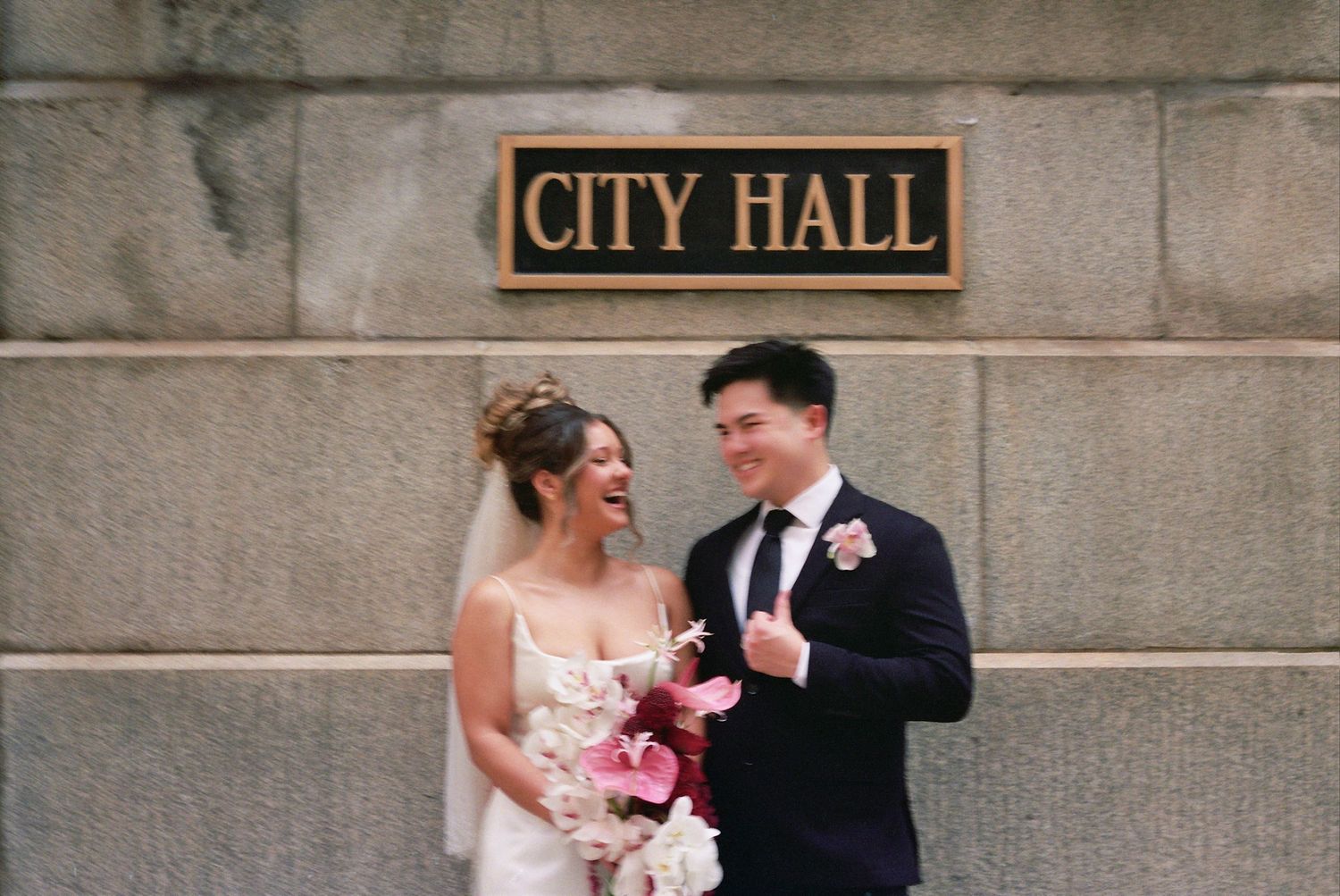 A newlywed couple shares a joyful moment outside a historic City Hall building with stone architecture.