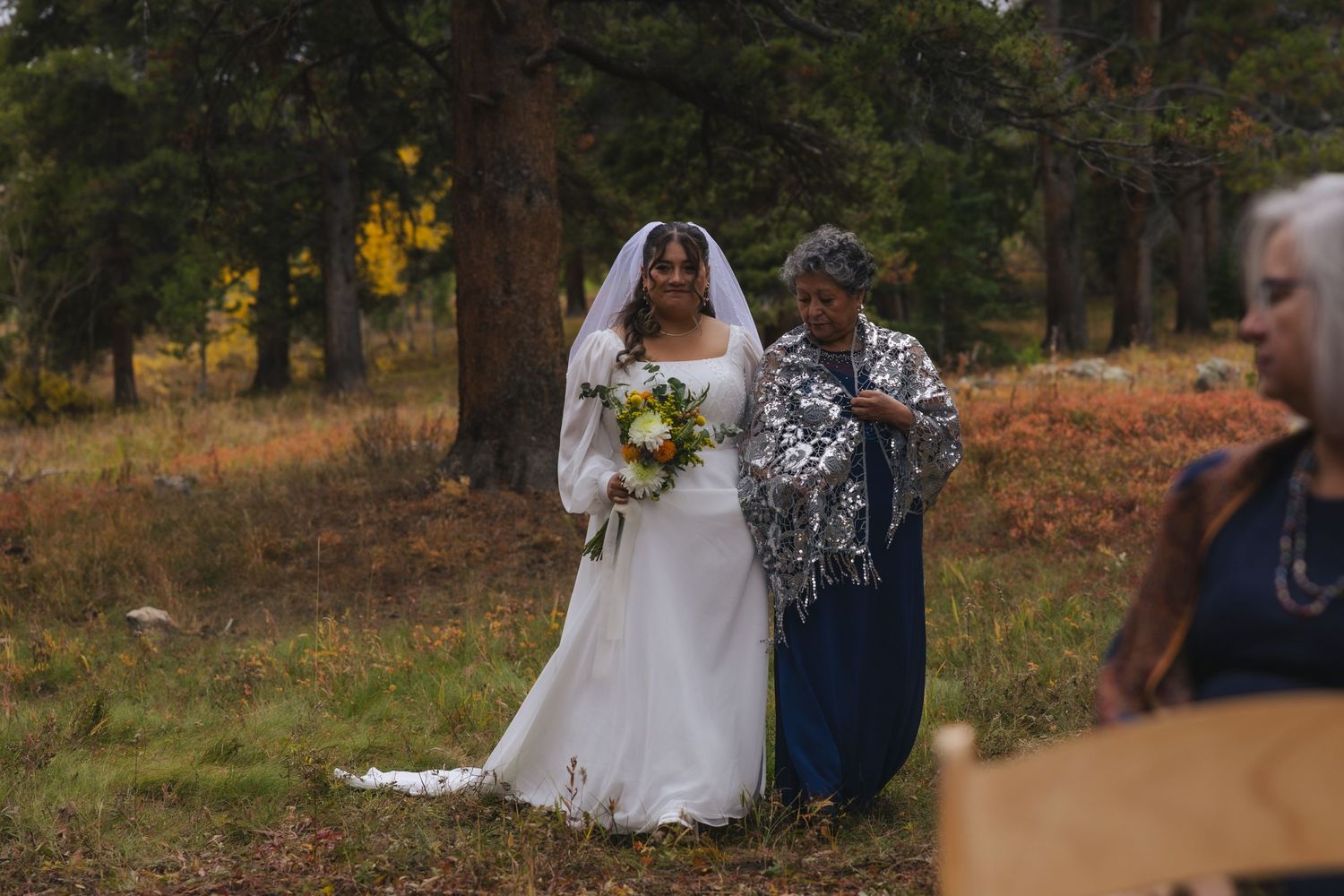 A bride in a white wedding dress holding a bouquet stands with a companion in blue outdoors during an autumn ceremony.