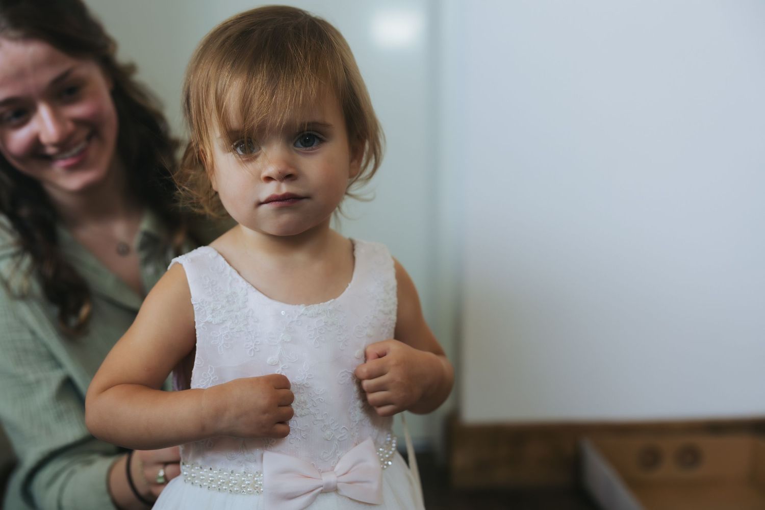 A small child wearing a white sleeveless dress stands indoors while someone smiles in the background.