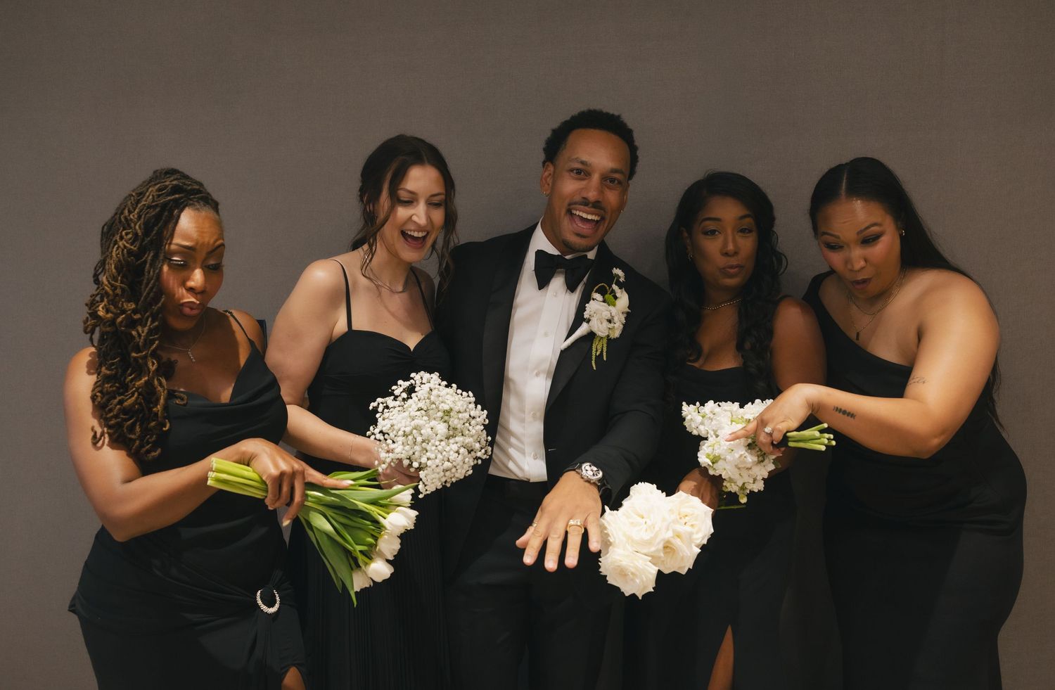 Groom in tuxedo poses with four bridesmaids in black dresses holding white bouquets against gray backdrop.