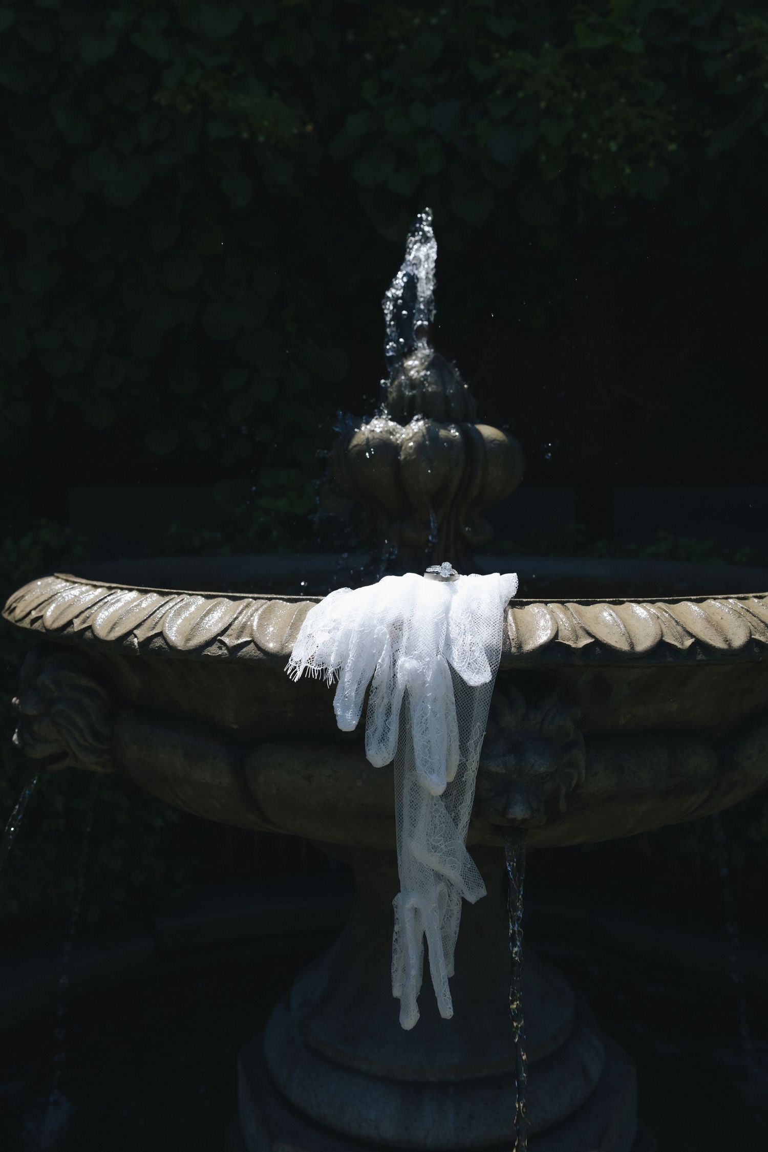 White gloves draped over an ornate stone garden fountain with water jet, surrounded by dark foliage.