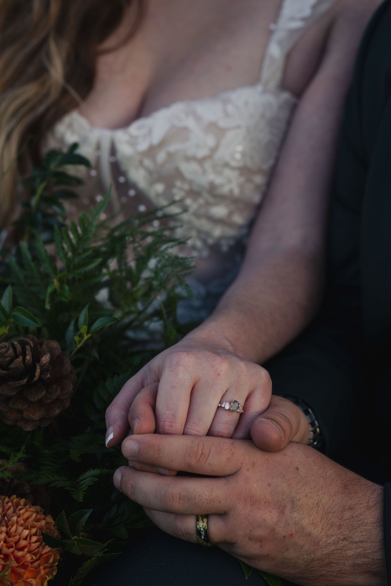Close-up of two hands with wedding rings intertwined next to greenery and pinecones in a romantic setting.