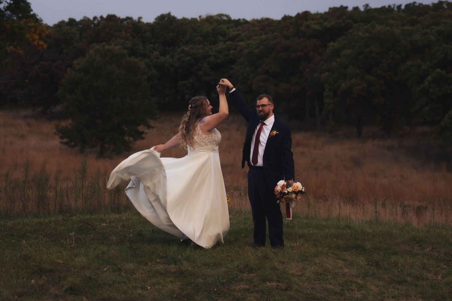 Wedding couple poses playfully in a rustic field as bride's dress catches the wind.