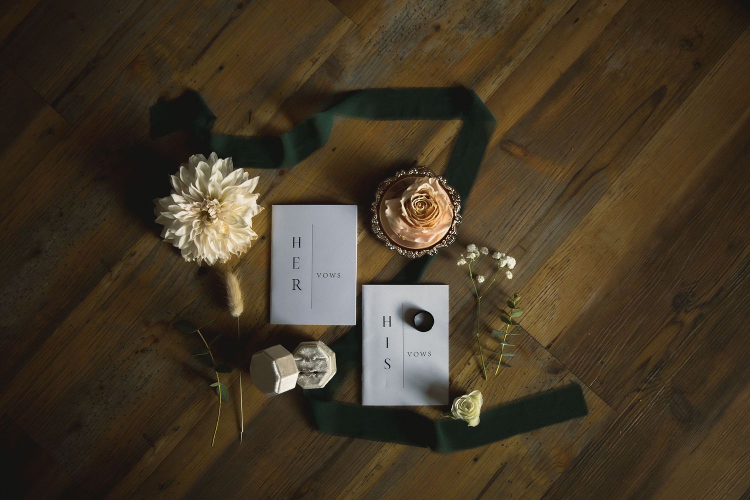 Wedding details flat lay with rings, dried flowers and cards on dark wood surface.