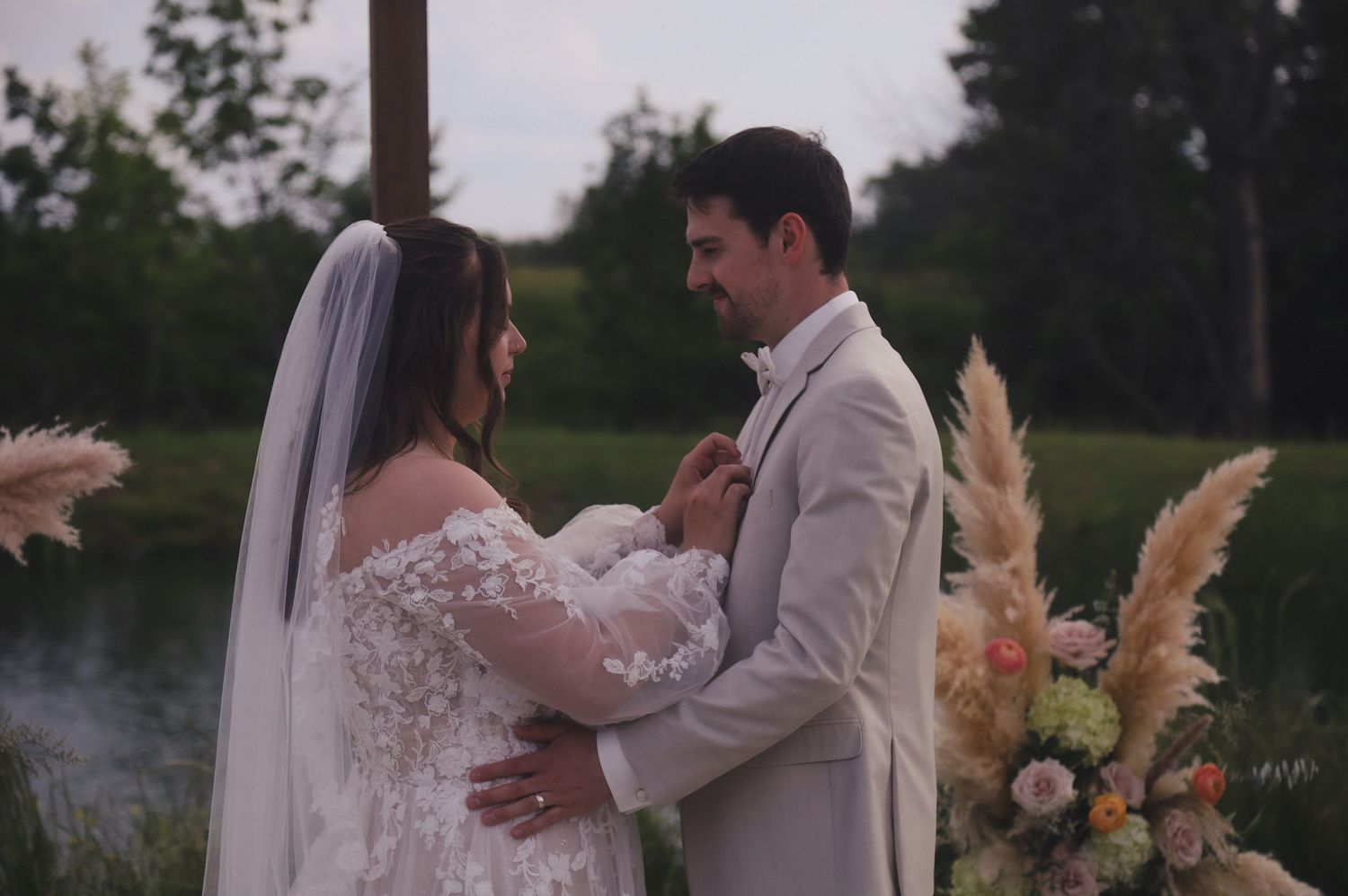 A couple embraces during their waterfront wedding ceremony with pampas grass floral arrangements in the background.