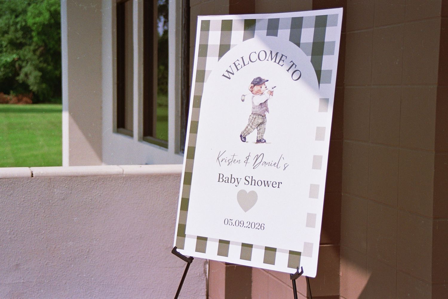 Welcome sign on easel for Regina and David's Baby Shower, featuring a golfer illustration on checkered background.