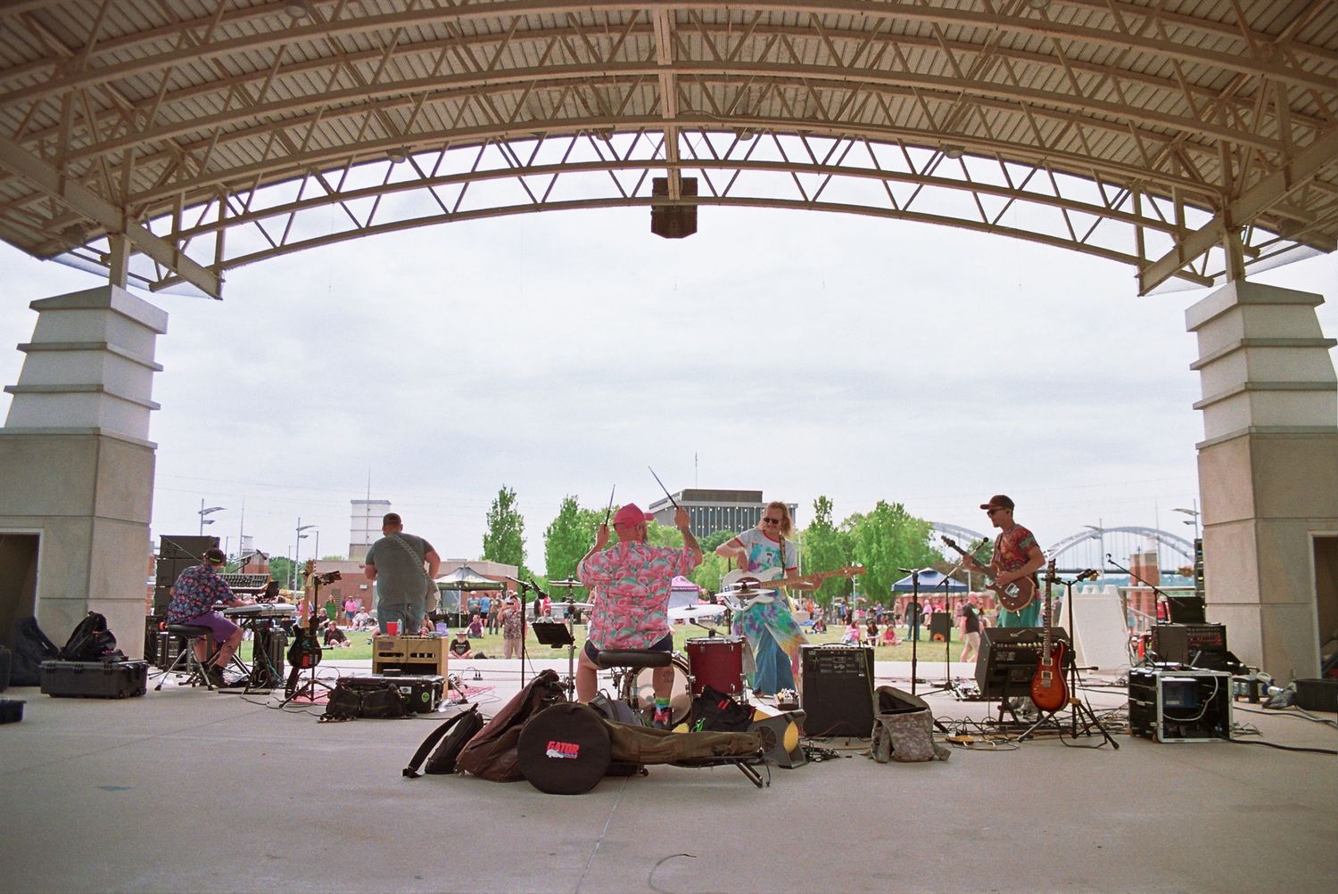 Band performing on an outdoor covered stage at a park amphitheater on a cloudy day.