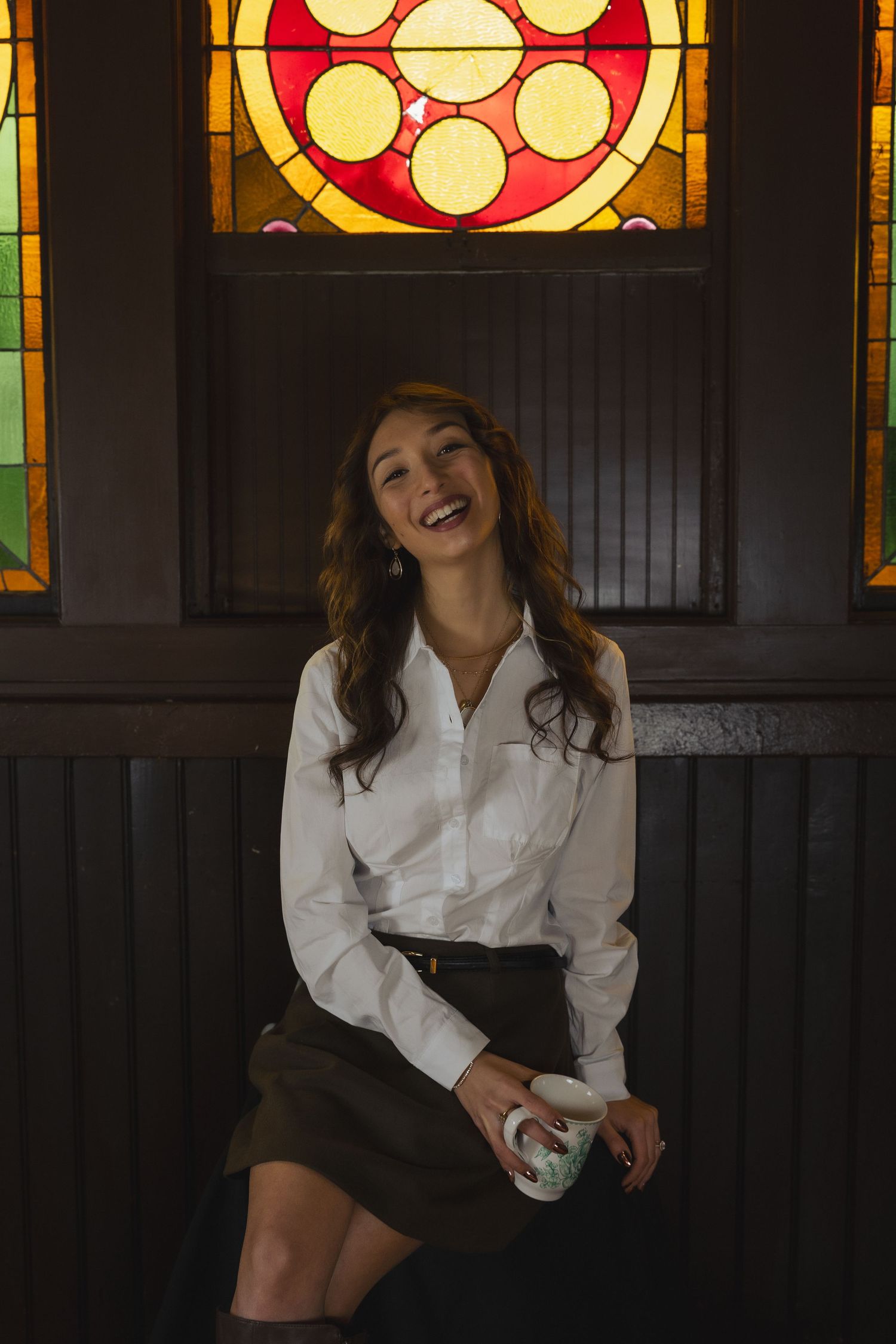 Smiling woman in white shirt and dark skirt sits holding a cup before a colorful stained glass window.