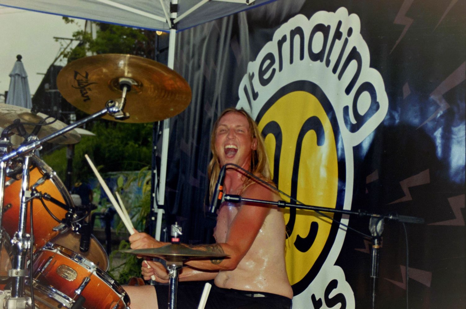 Energetic female drummer performing shirtless on outdoor stage in front of Alternating Current festival banner.