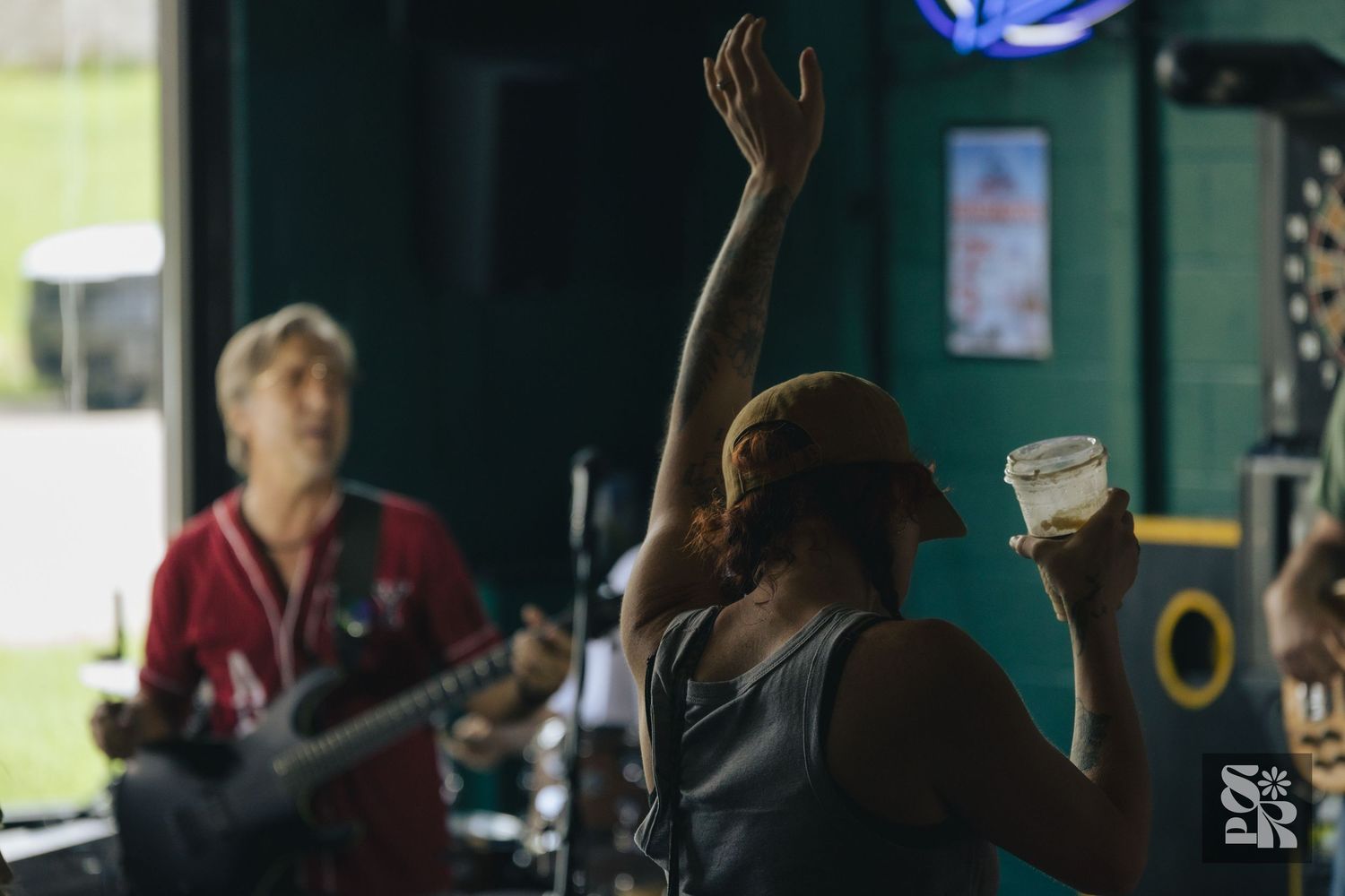 A person raises their hand holding a drink at a live music venue while a guitarist performs in the background.
