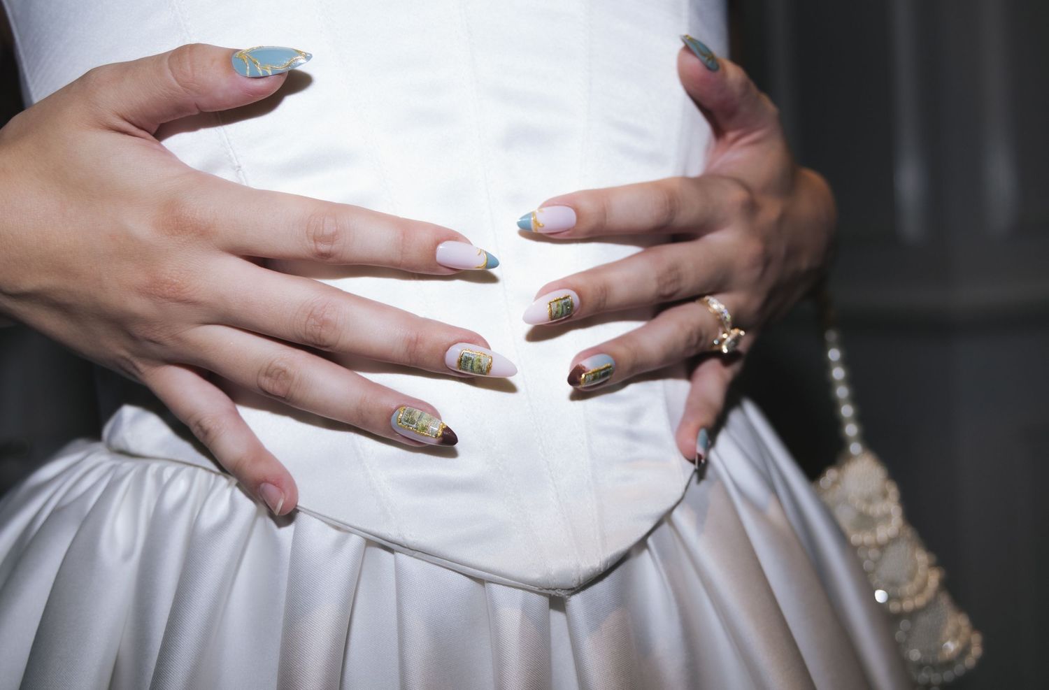 Close-up of hands with long stiletto nails featuring pastel nail art, resting on a white satin dress.