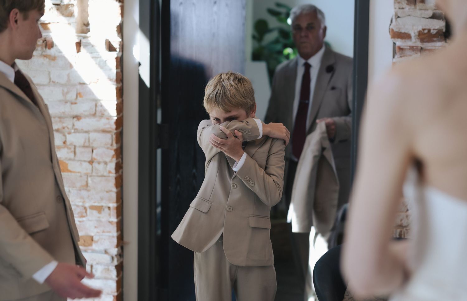 Young boy in beige suit covering face at formal event with adults in background near brick wall doorway.