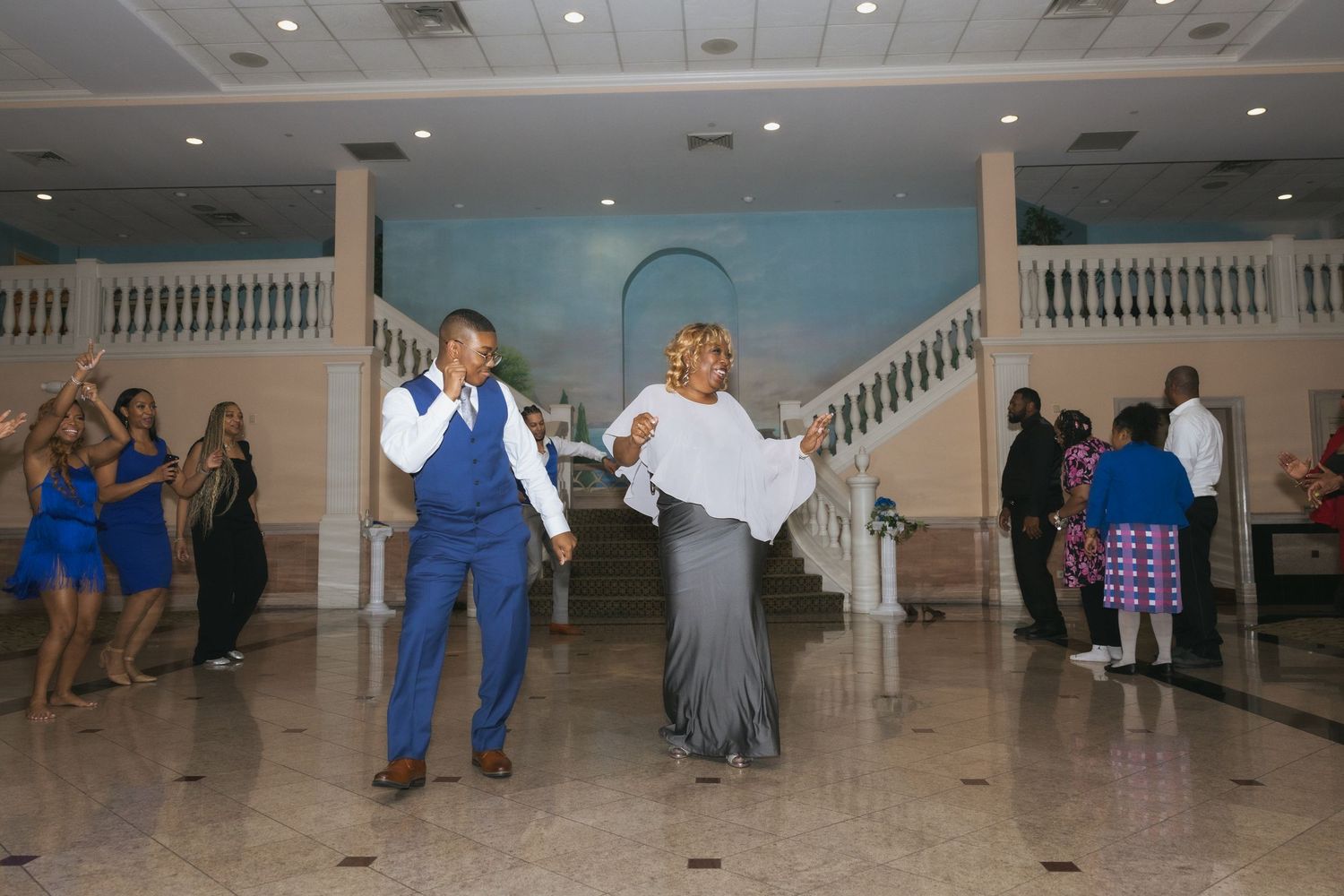 Couple dancing joyfully at a wedding reception in an elegant ballroom with guests in blue attire.