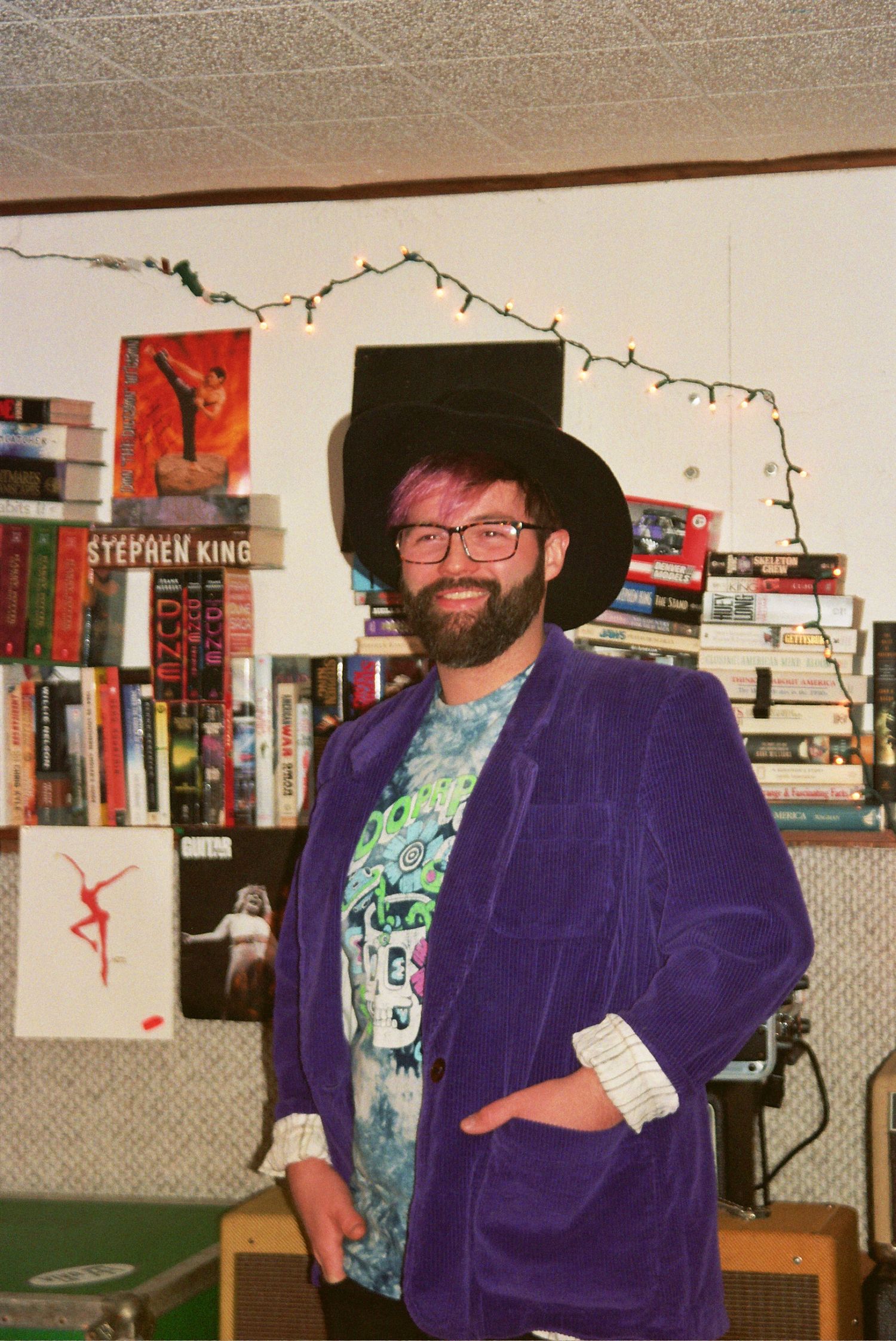 Bearded man in purple velvet blazer and black hat smiling in front of a bookshelf with Stephen King books.