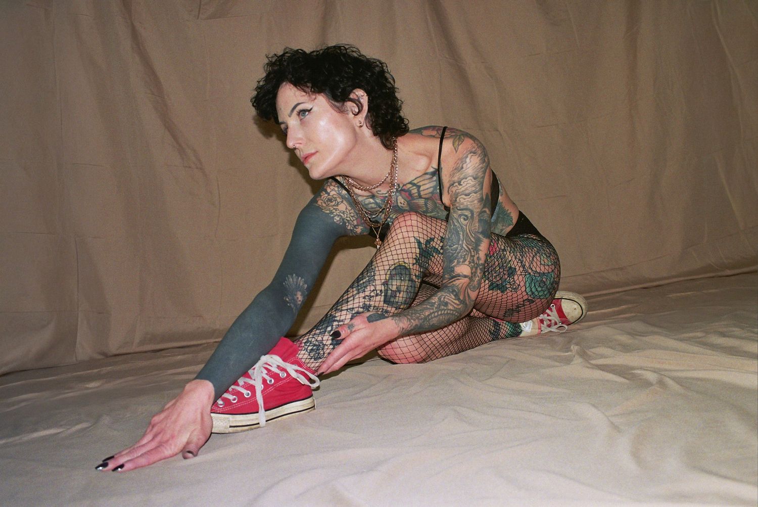 Heavily tattooed woman in fishnet bodysuit and red Converse sneakers posing on white floor against beige backdrop.