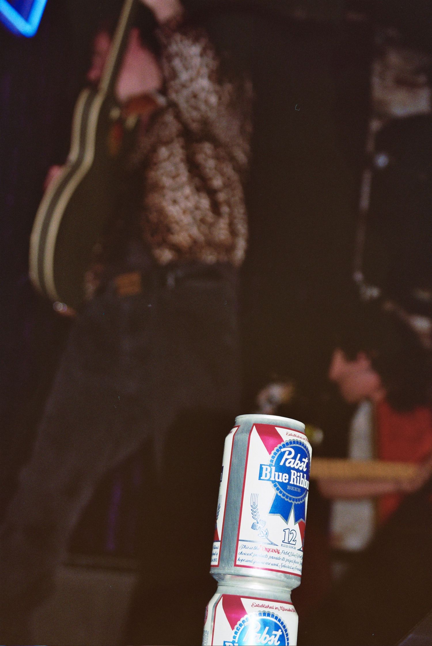 A Pabst Blue Ribbon beer can on a reflective surface at a live music venue with band performing in background.