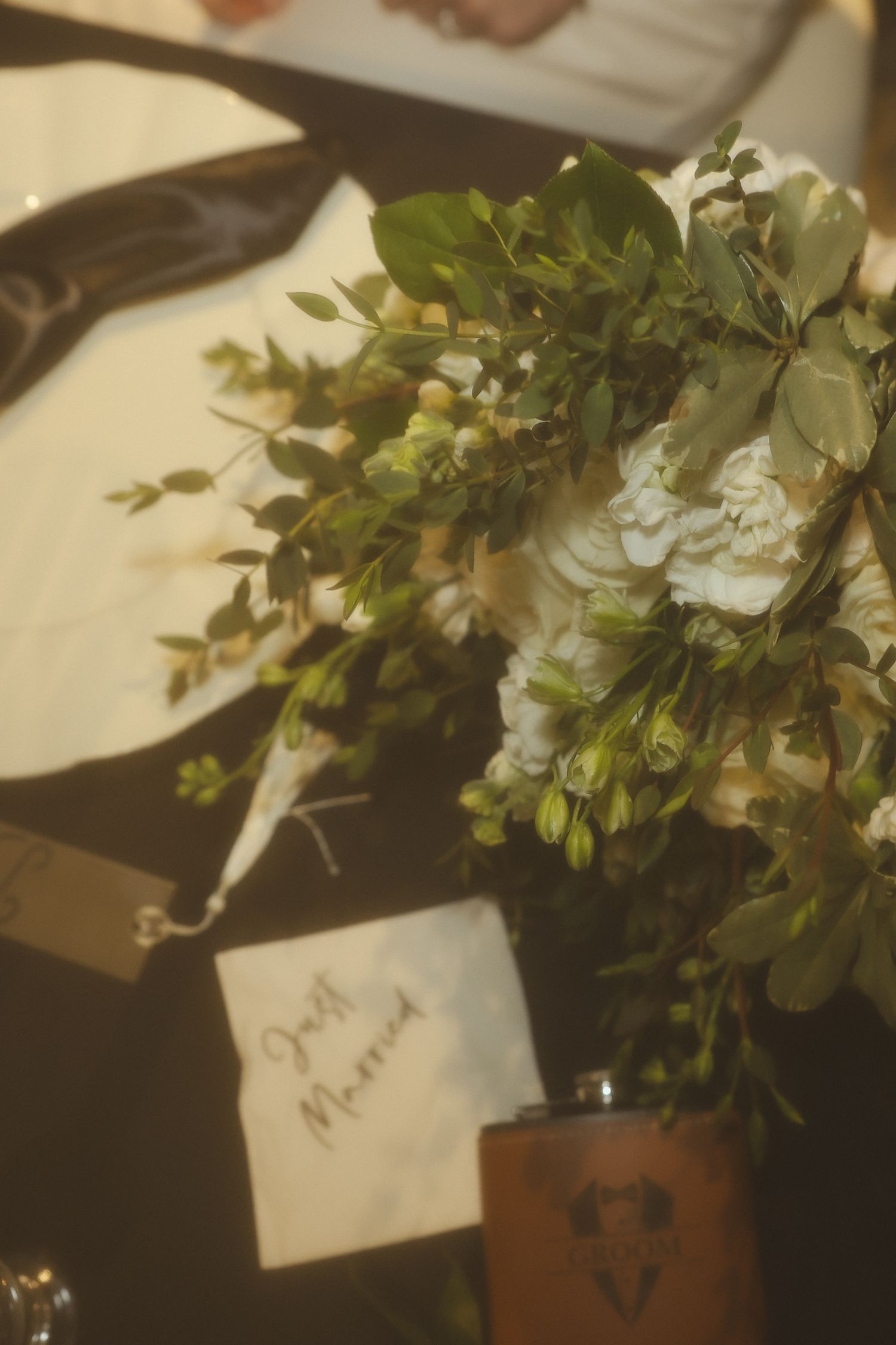 A small note card sits near a flower arrangement with white blooms and green foliage in a brown vase.