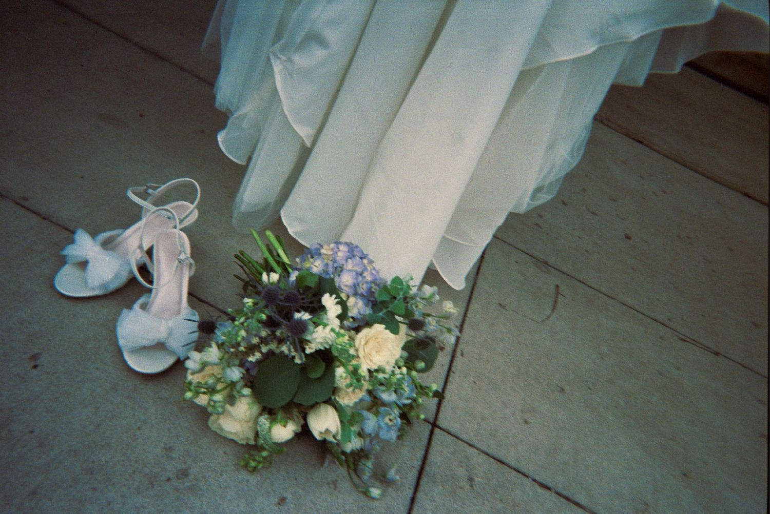 A white bridal bouquet with blue accents and white wedding shoes on a concrete floor next to flowing fabric.