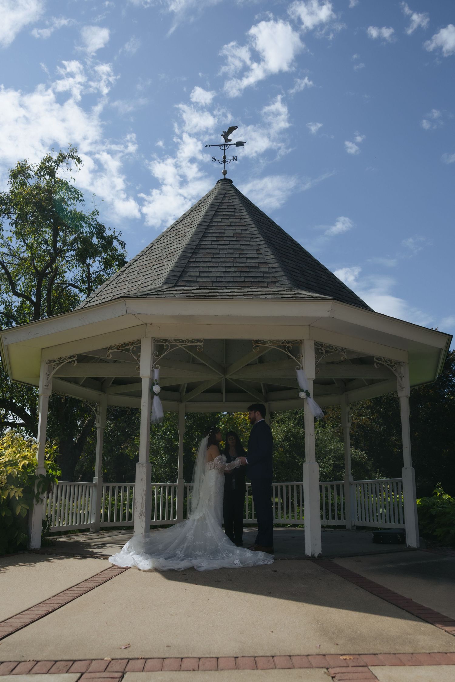 A white wedding gazebo with a hexagonal roof stands against a blue sky with puffy clouds on a sunny day.